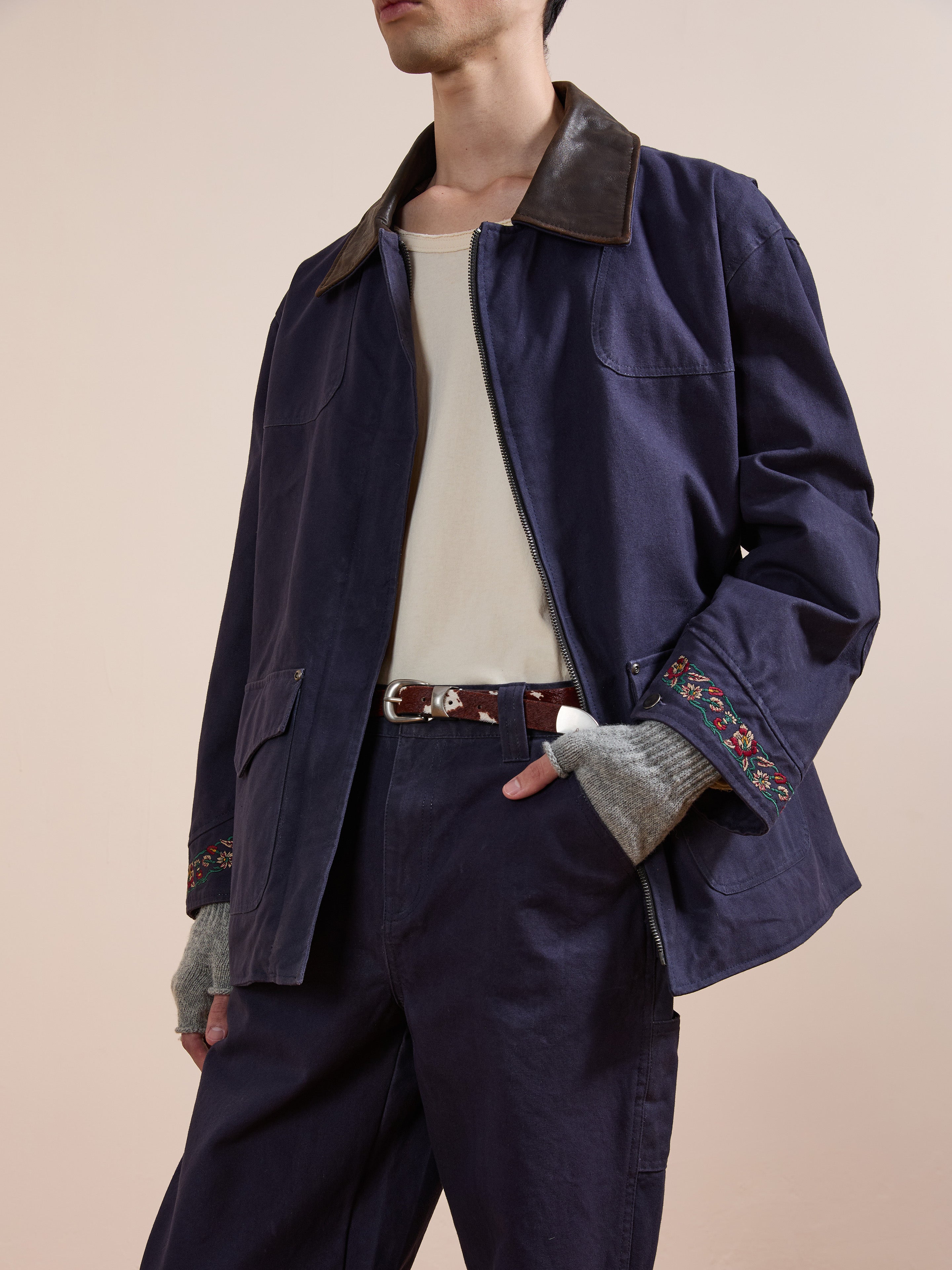 A person wears the Found Canvas Distressed Barn Jacket featuring a distressed leather collar and floral embroidery on the cuffs, paired with a cream shirt, dark pants, brown belt, and fingerless gray gloves against a beige background.