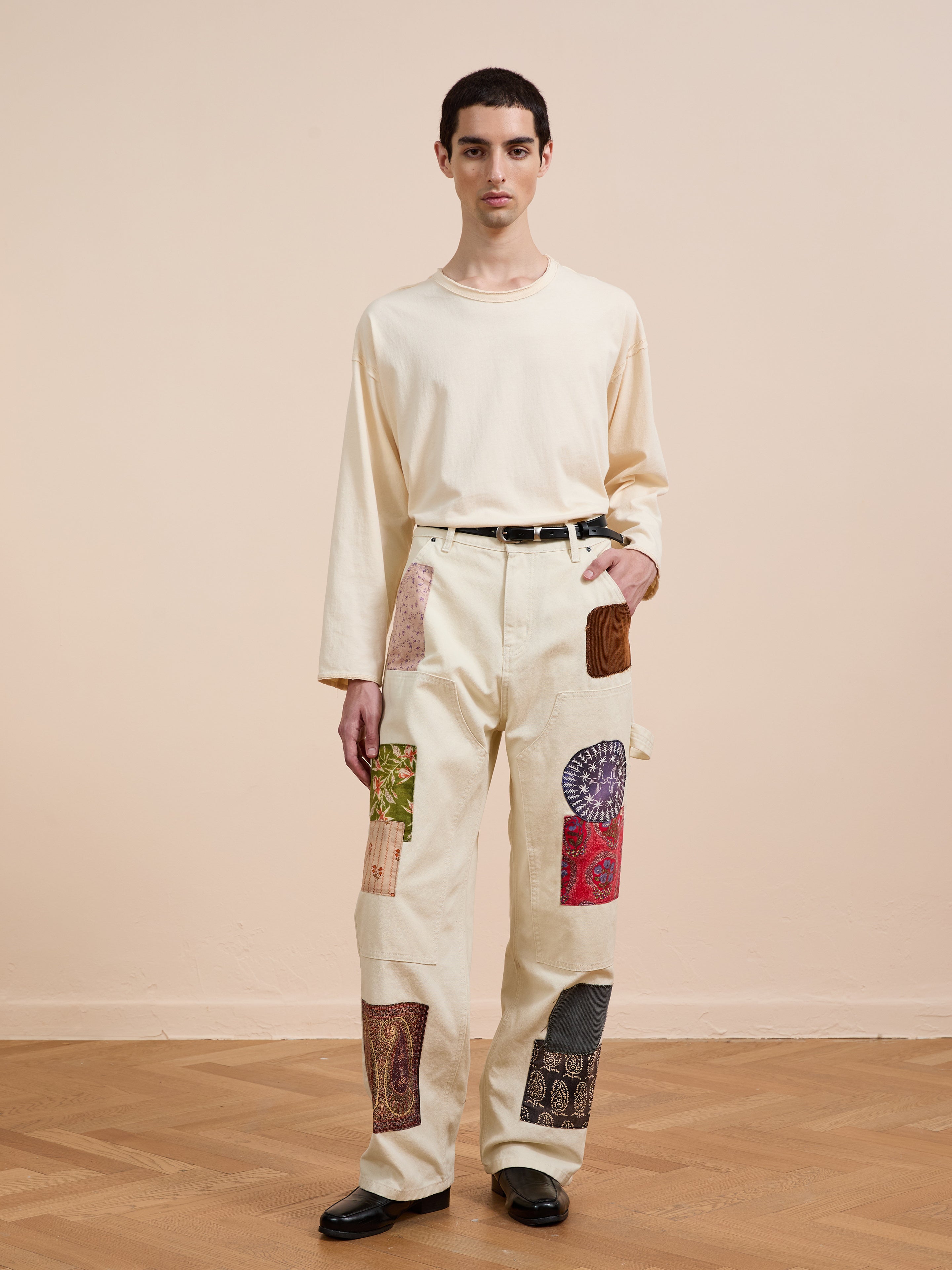 A person stands indoors on a wooden floor wearing FOUND's Tapestry Carpenter Canvas Pants—relaxed fit with multicolored Pakistani quilt-inspired patches—paired with a cream long-sleeve shirt, black belt, and black shoes.