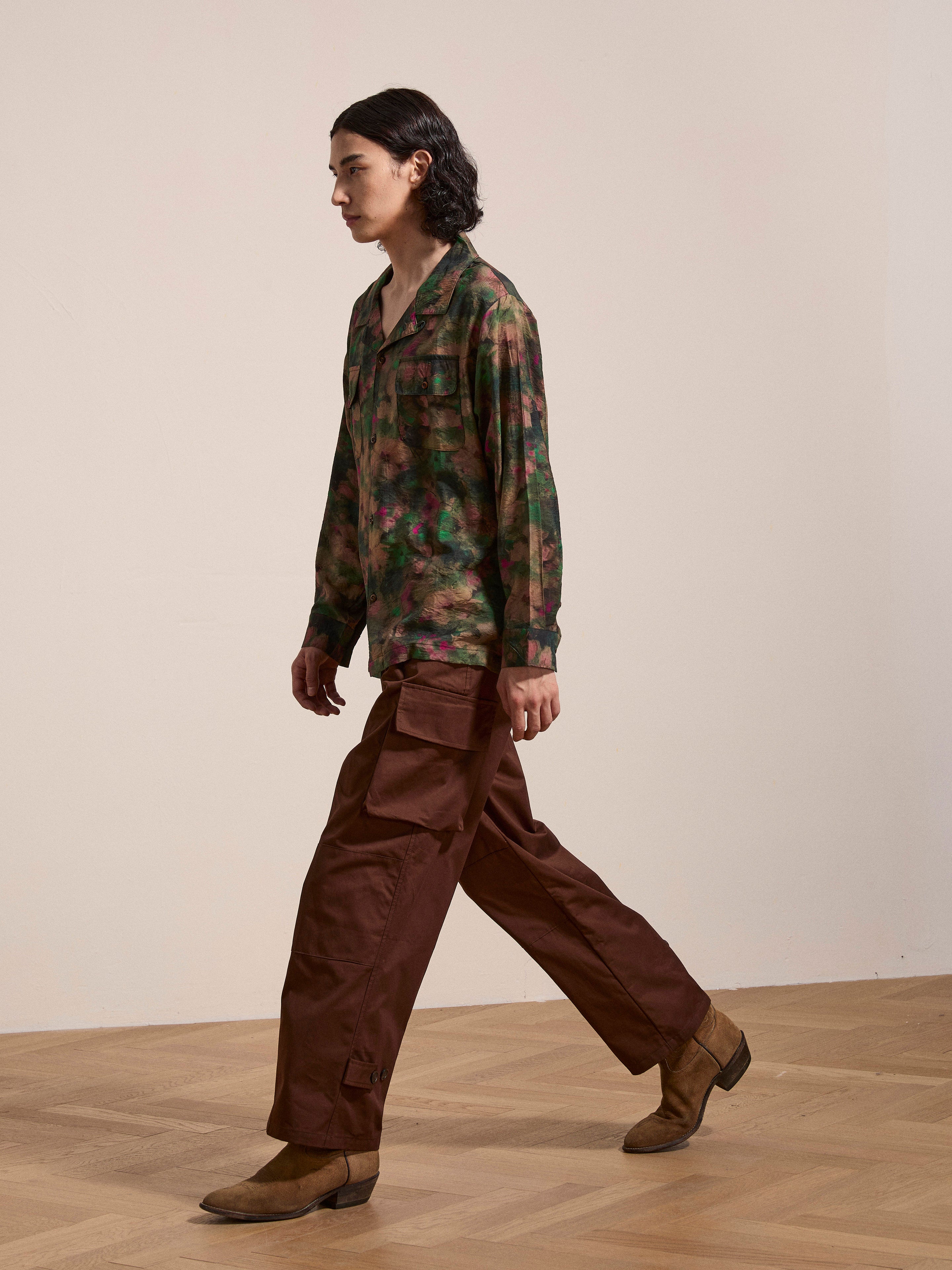Person walking indoors, dressed in a Charlot LS Camp Shirt from FOUND featuring a green camo pattern and dual chest pockets, brown cargo pants, and tan boots, with wavy dark hair.