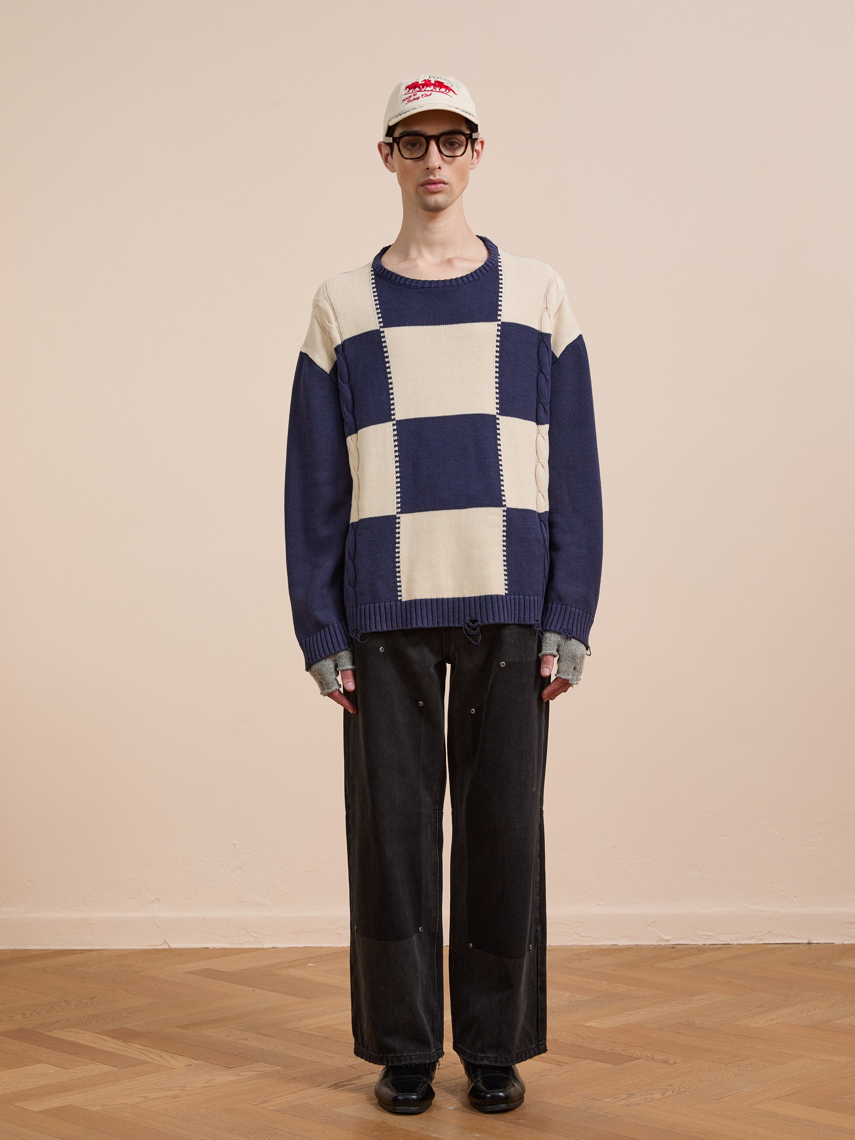 A person stands against a plain beige wall wearing the Found Checkered Fisherman Sweater in cream and navy, styled with relaxed fit, black wide-leg pants, black shoes, glasses, and a beige baseball cap.