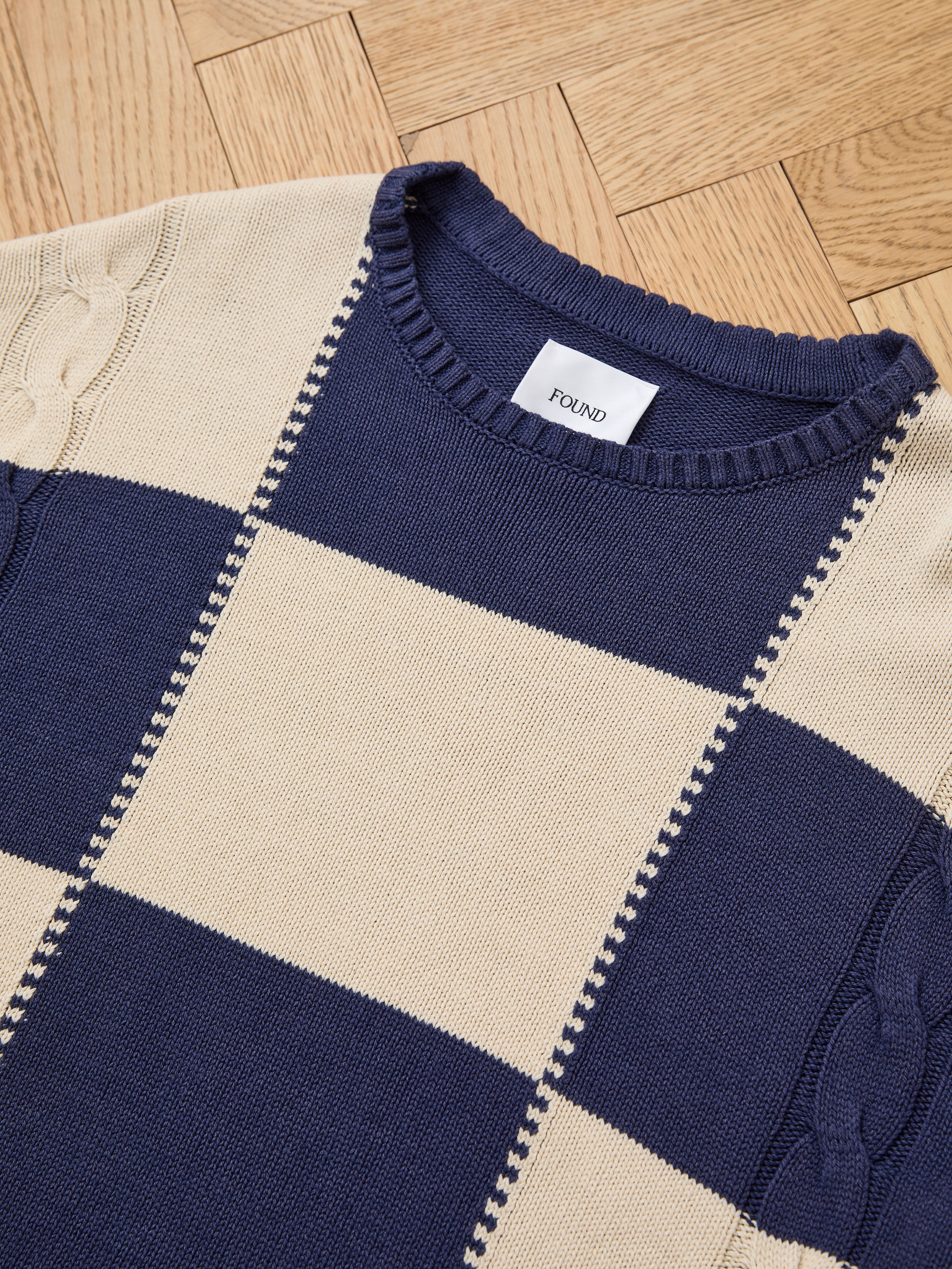 A checkered knit sweater from Found, featuring a mid-weight unisex relaxed fit, is laid flat on a herringbone wood floor.
