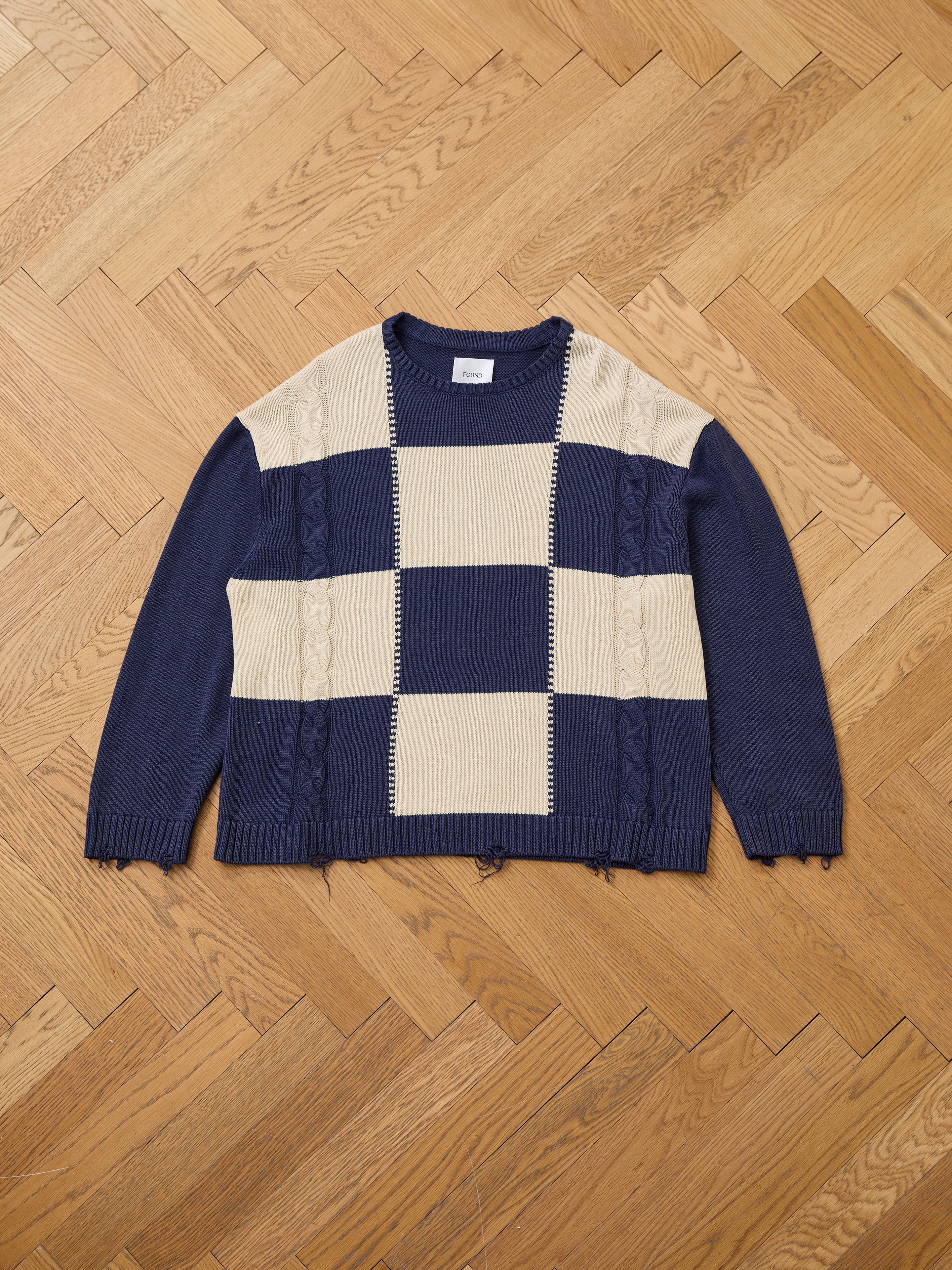 The Found Checkered Fisherman Sweater in navy blue and beige features a crew neck, cable knit details, ribbed cuffs, and a relaxed unisex fit. This mid-weight sweater offers true-to-size comfort for effortless everyday style.