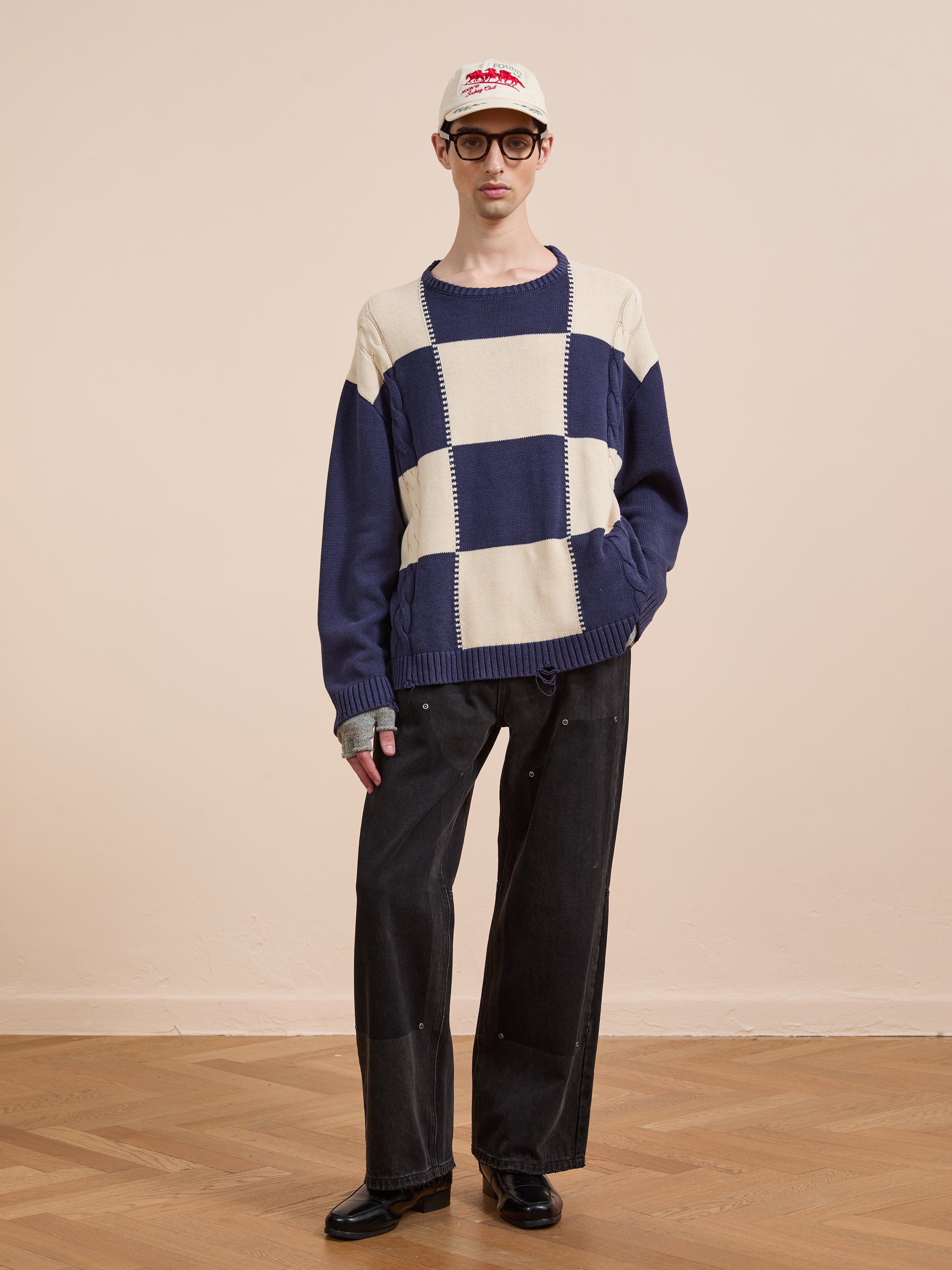 A person wears the Found Checkered Fisherman Sweater, styled with black wide-leg jeans, black shoes, glasses, and a white cap, standing against a plain beige wall on a wooden floor.