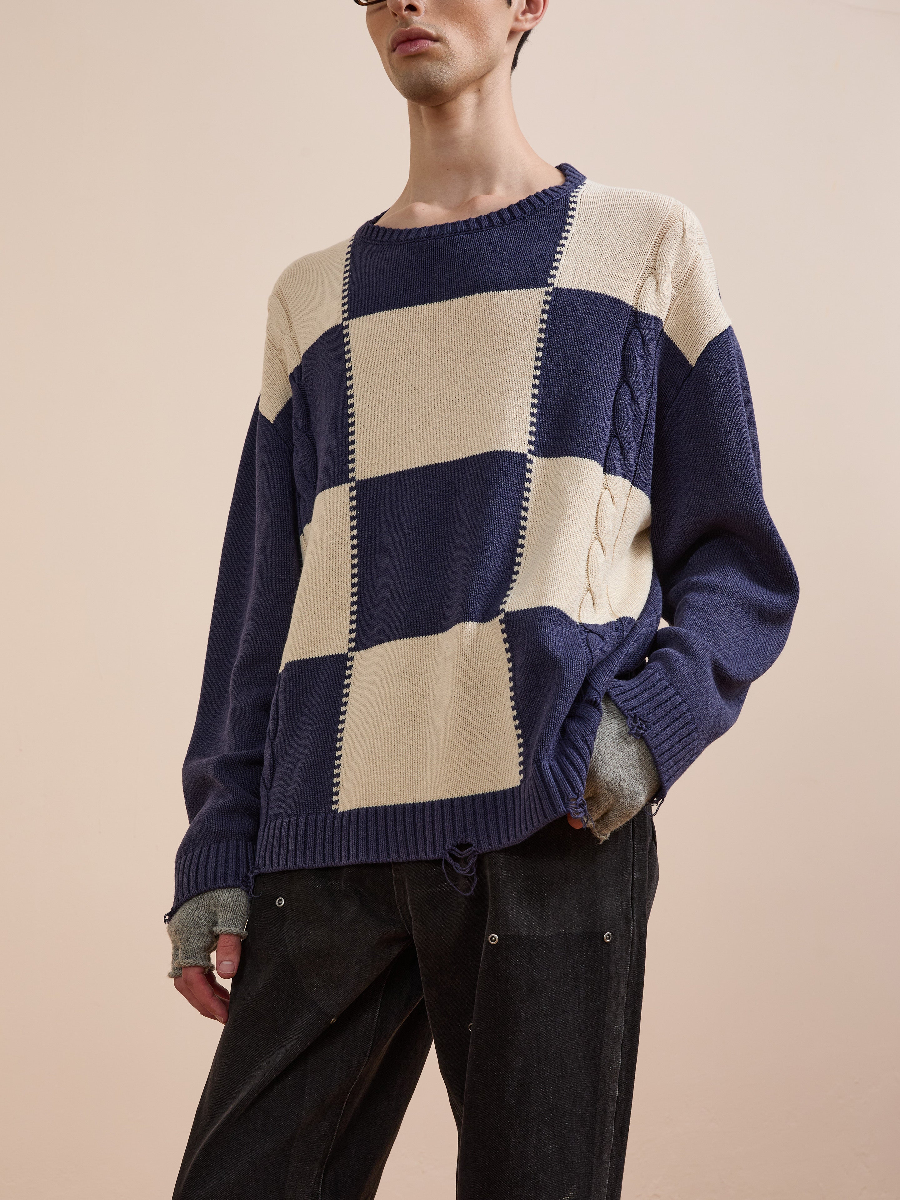 Wearing Found’s Checkered Fisherman Sweater in navy and beige, styled with gray fingerless gloves and black pants, a person stands against a plain beige background.