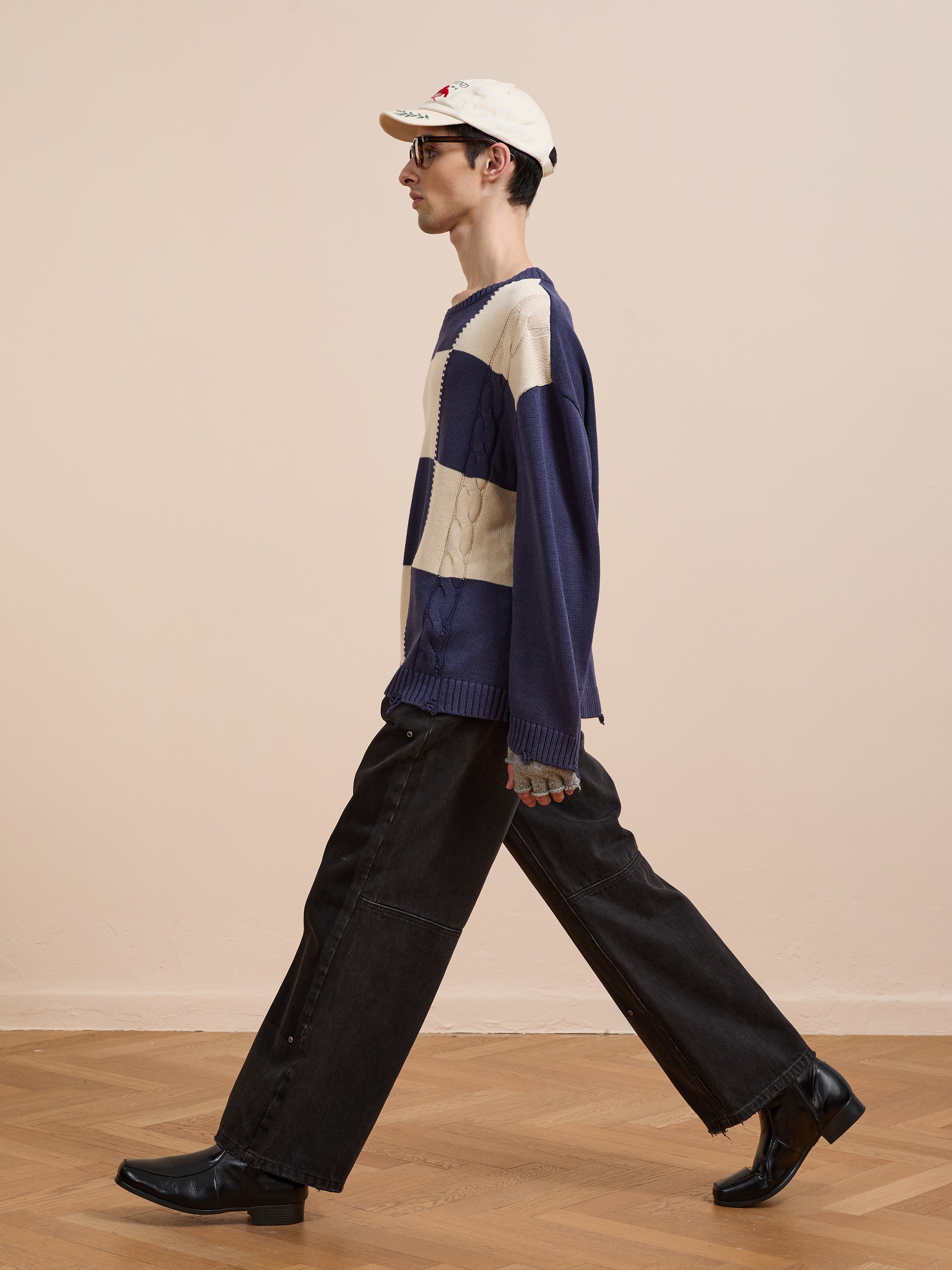 A person wearing Found's Checkered Fisherman Sweater, a cream baseball cap, sunglasses, wide-leg black jeans, and black heeled boots walks indoors on a wooden floor.