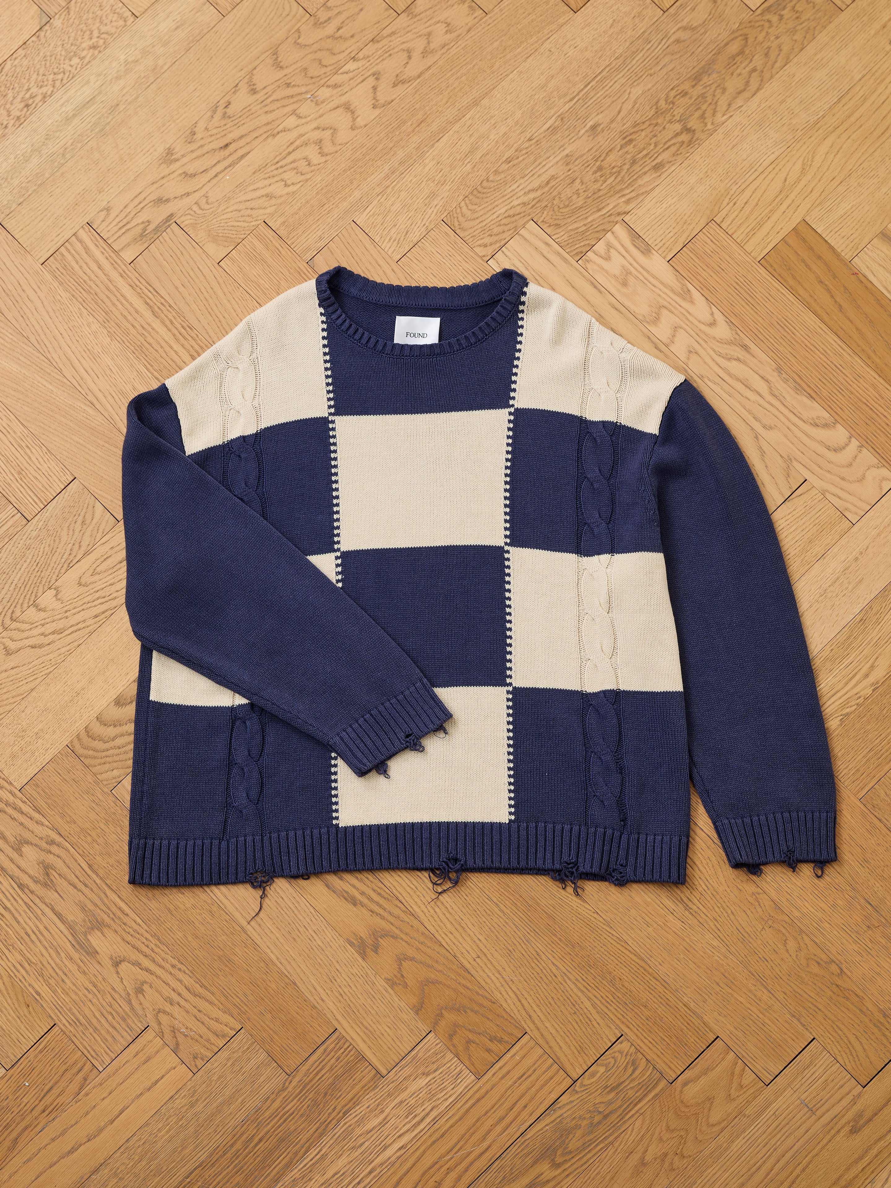 The Found Checkered Fisherman Sweater, a unisex navy and beige long-sleeve with wide stripes and cable knit details, features a relaxed fit that's true to size and is shown laid flat on a light wood herringbone floor.