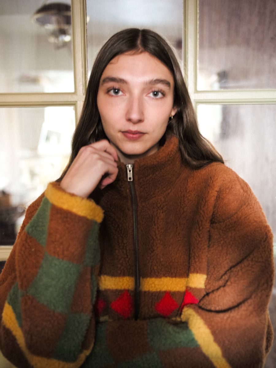 A person with long hair, wearing a Checkered Zip-Up Sherpa by FOUND, stands in front of a glass-paneled door, looking directly at the camera.