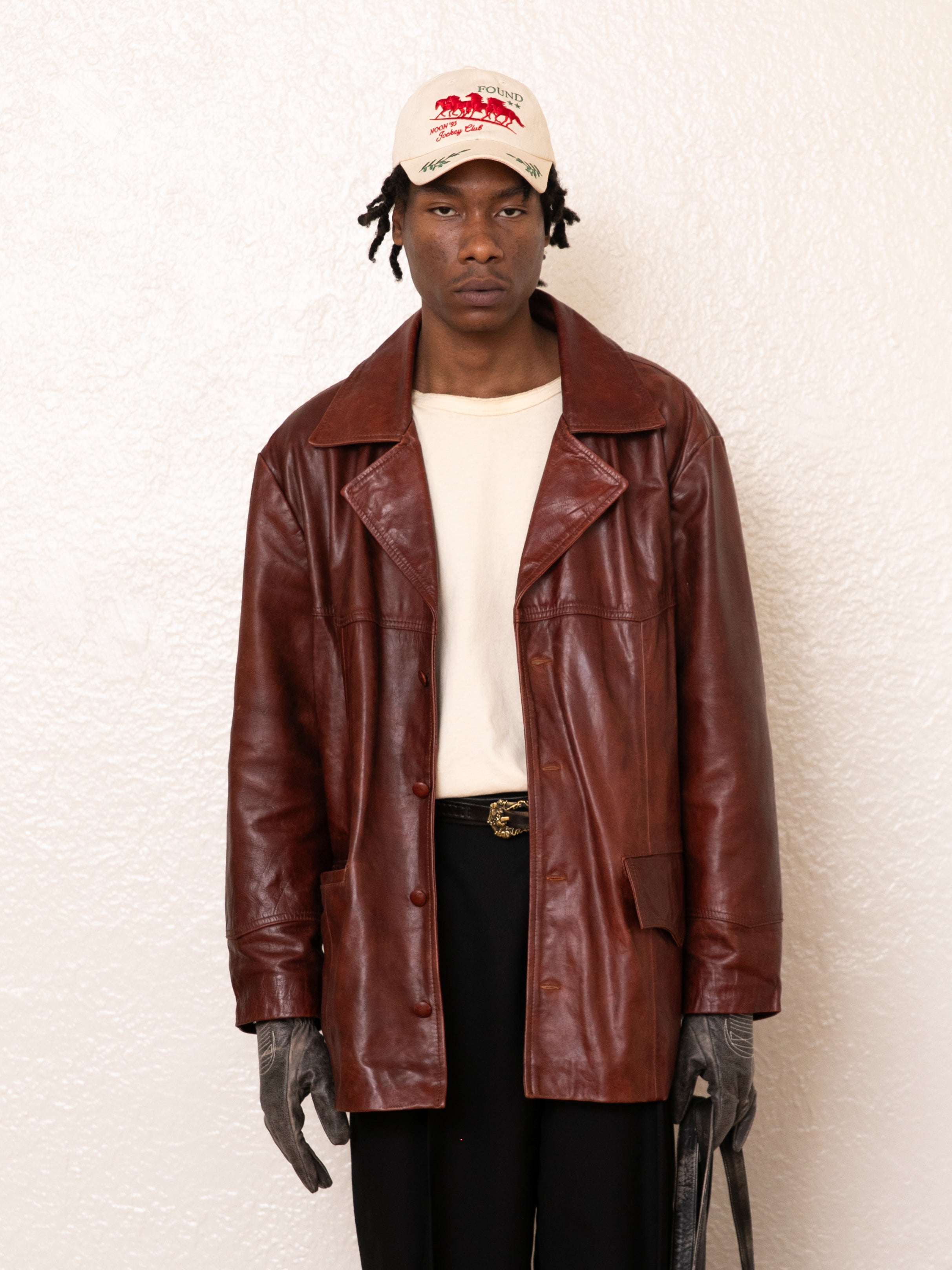 Against a white background, an individual wears the FOUND Claret Western Oversized Leather Blazer over a white shirt, paired with black pants and gray gloves. They finish the look with a white cap featuring red text for an effortlessly stylish ensemble.