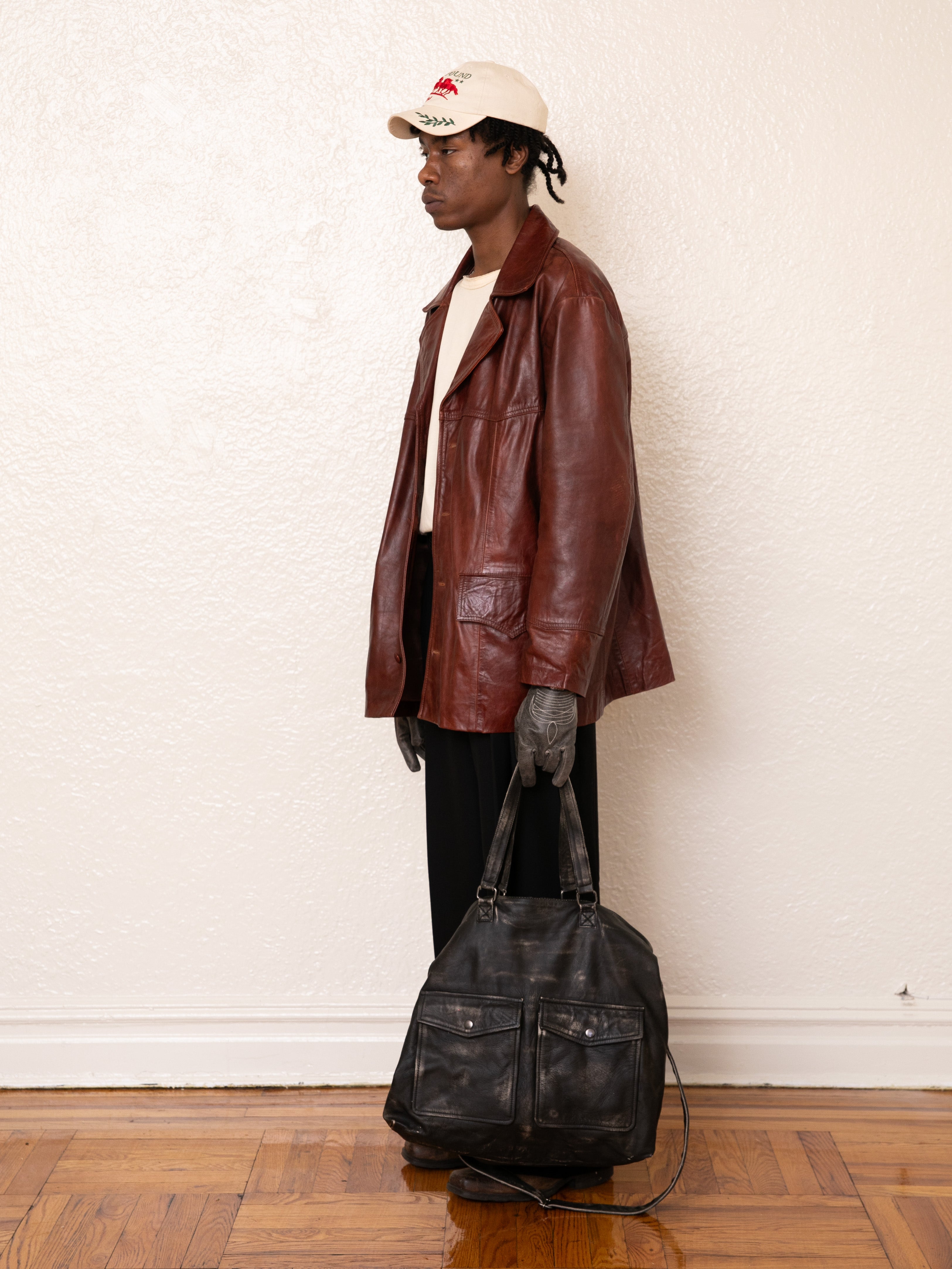 A person wearing a FOUND Claret Western Oversized Leather Blazer, cream cap, and black pants holds a large black bag, standing against a white wall on a wooden floor.