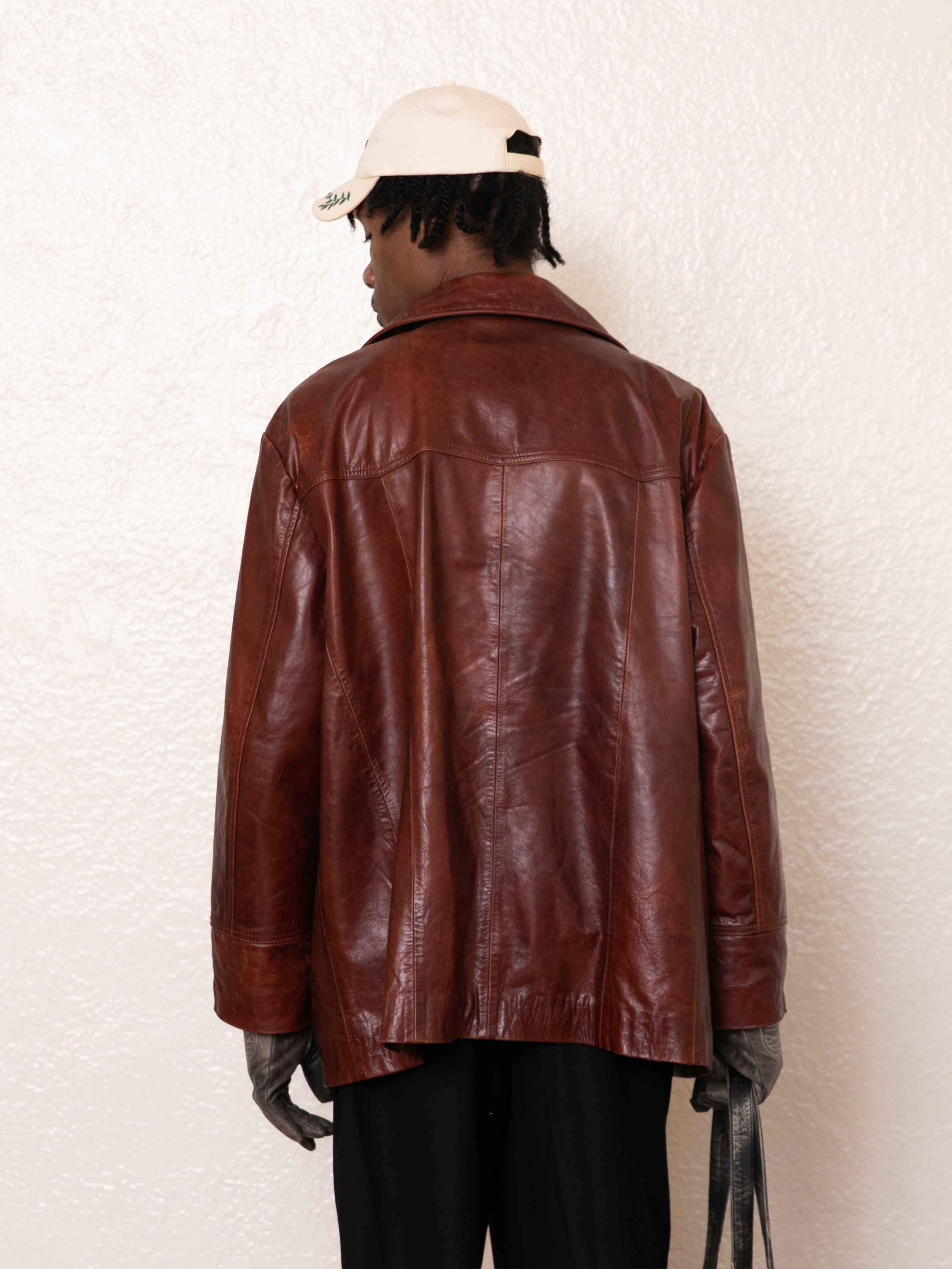 A person in a claret Western oversized leather blazer by FOUND, paired with black pants, gloves, and a light-colored cap, stands facing away against a textured white background.
