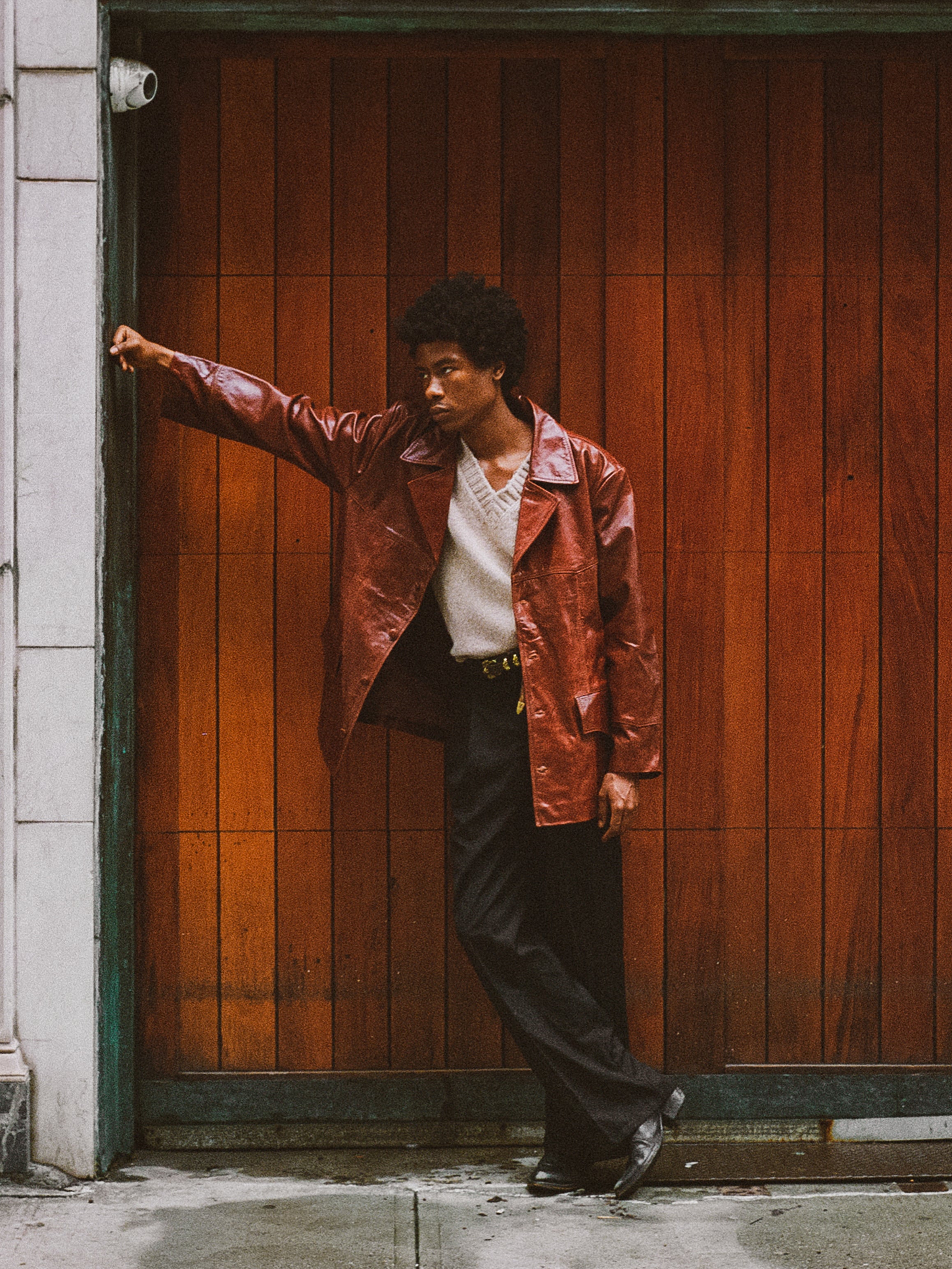 A person in a claret western oversized leather blazer by FOUND and black pants leans against a wooden door, one arm extended.