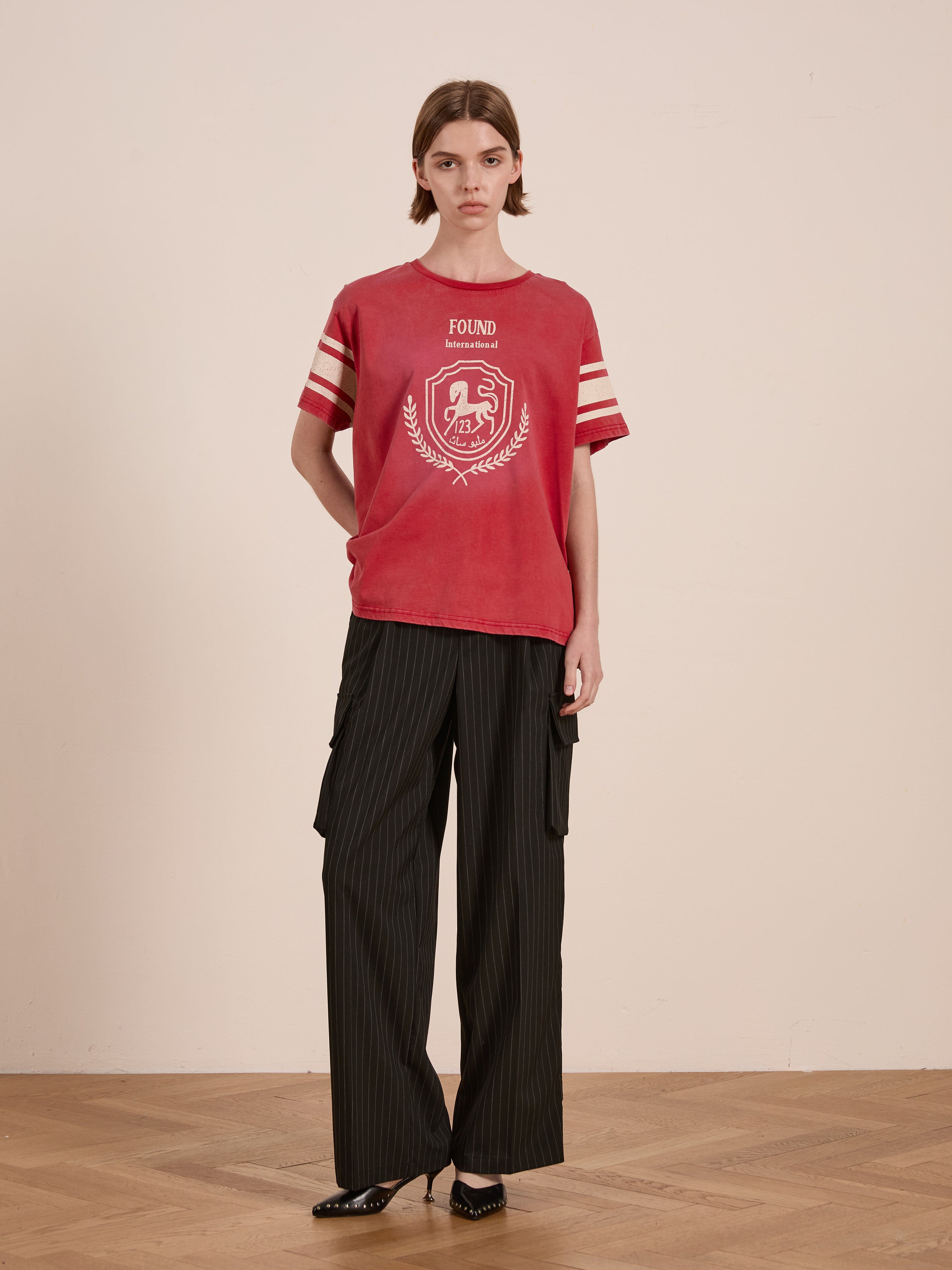 A person wearing a FOUND Collegiate Crest Tee in red and black pinstripe cargo pants stands on a wooden floor against a plain beige background.
