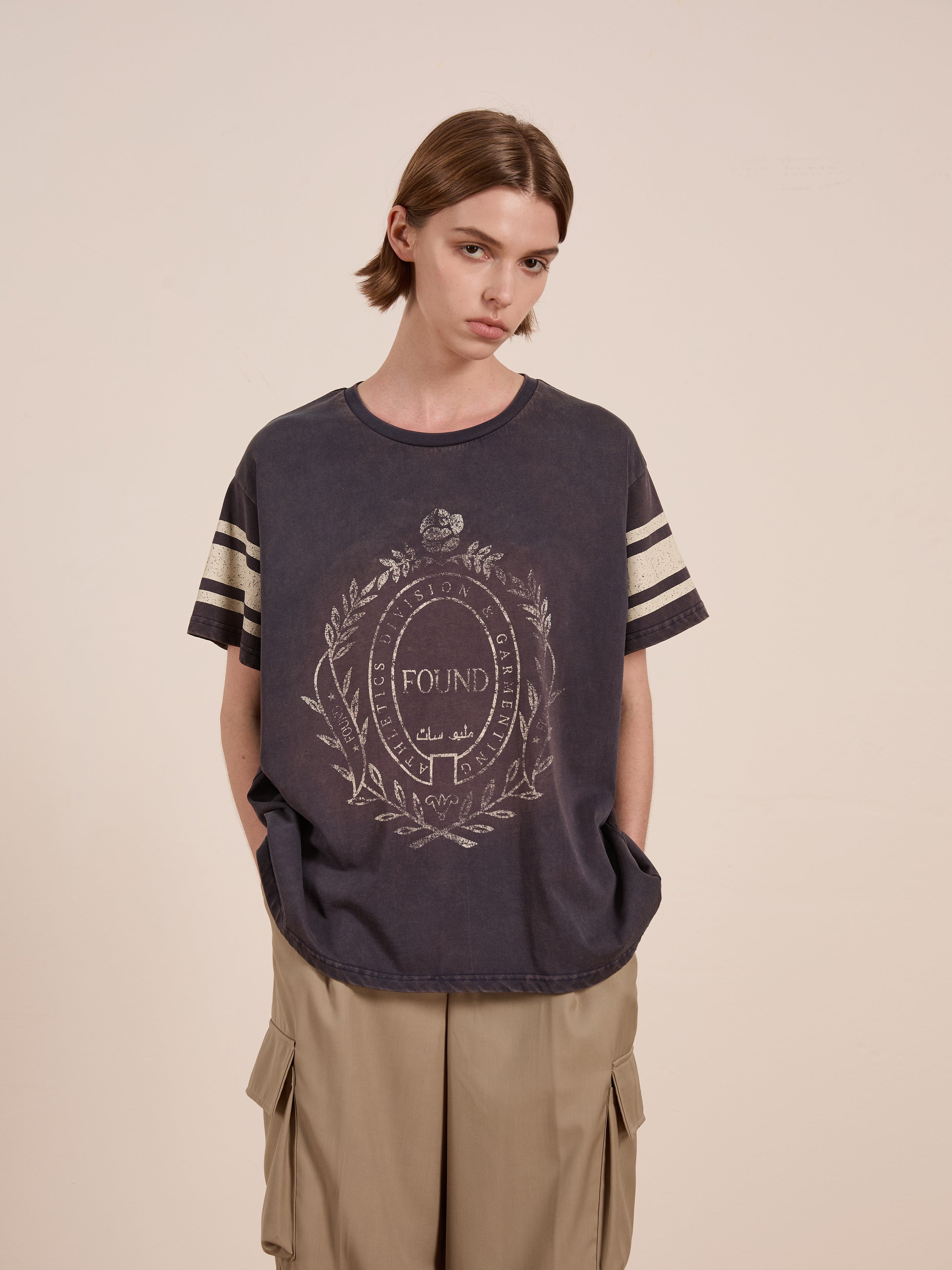 A person in a black Collegiate Crest Tee by FOUND, paired with tan cargo pants against a plain background. The unisex relaxed fit tee adds vintage athletic charm to the outfit.