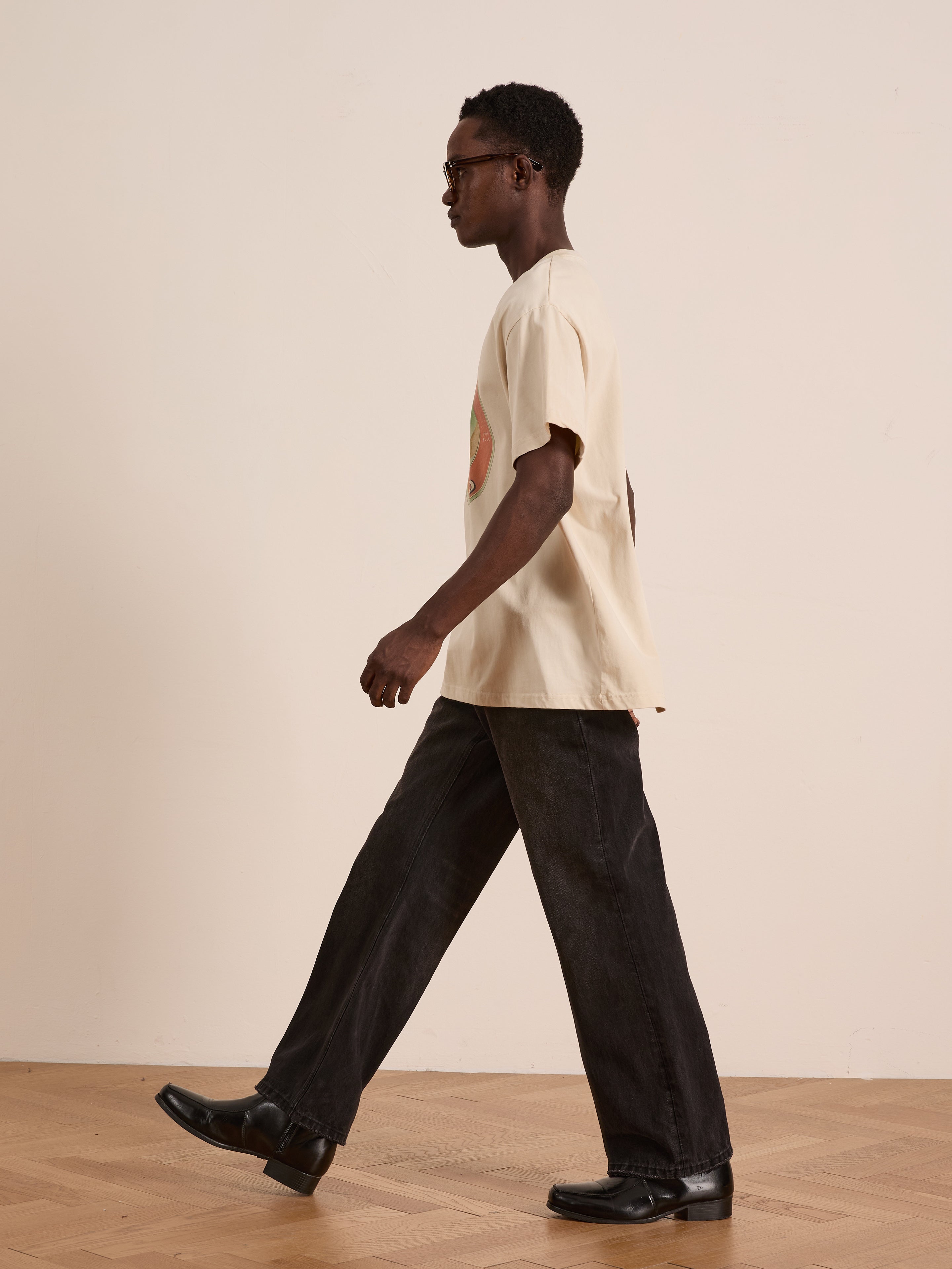 A man wearing the FOUND Community Hands Tee, black jeans, and black shoes walks indoors on a wooden floor against a plain light-colored wall.