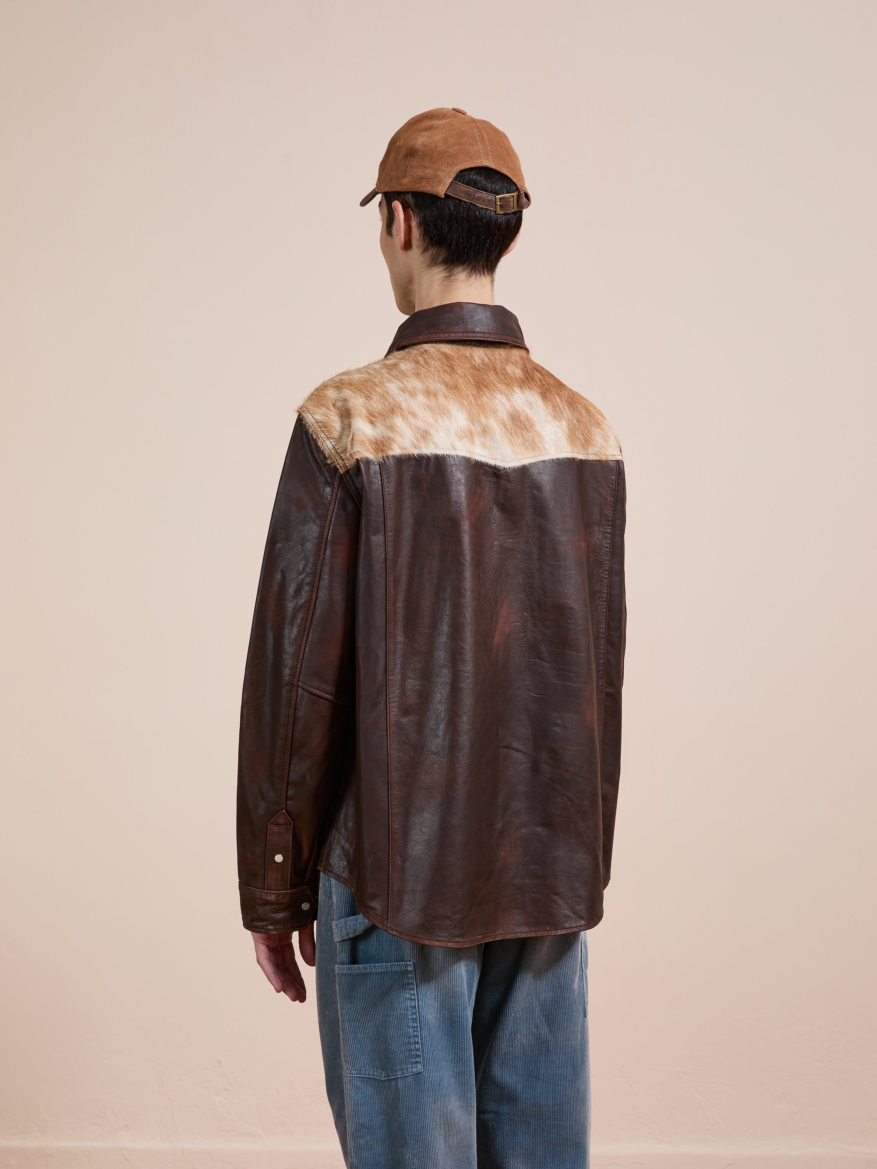A person in the FOUND Distressed Leather Cowhide Contrast Western Shirt, blue jeans, and a brown cap stands with their back to the camera against a plain background.