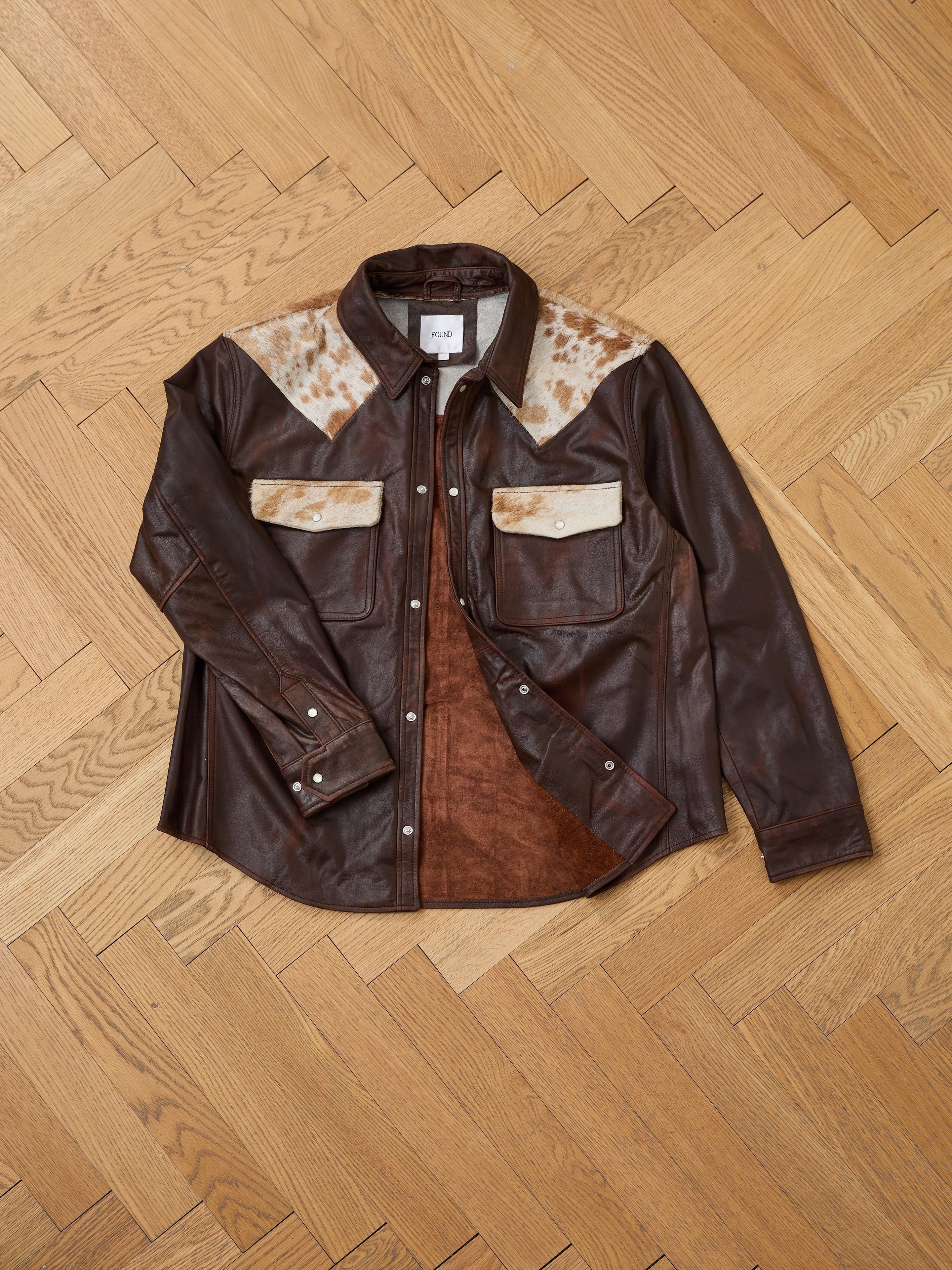 The FOUND Distressed Leather Cowhide Contrast Western Shirt, featuring white and brown patches on the shoulders and pockets, is laid flat on a wooden floor.