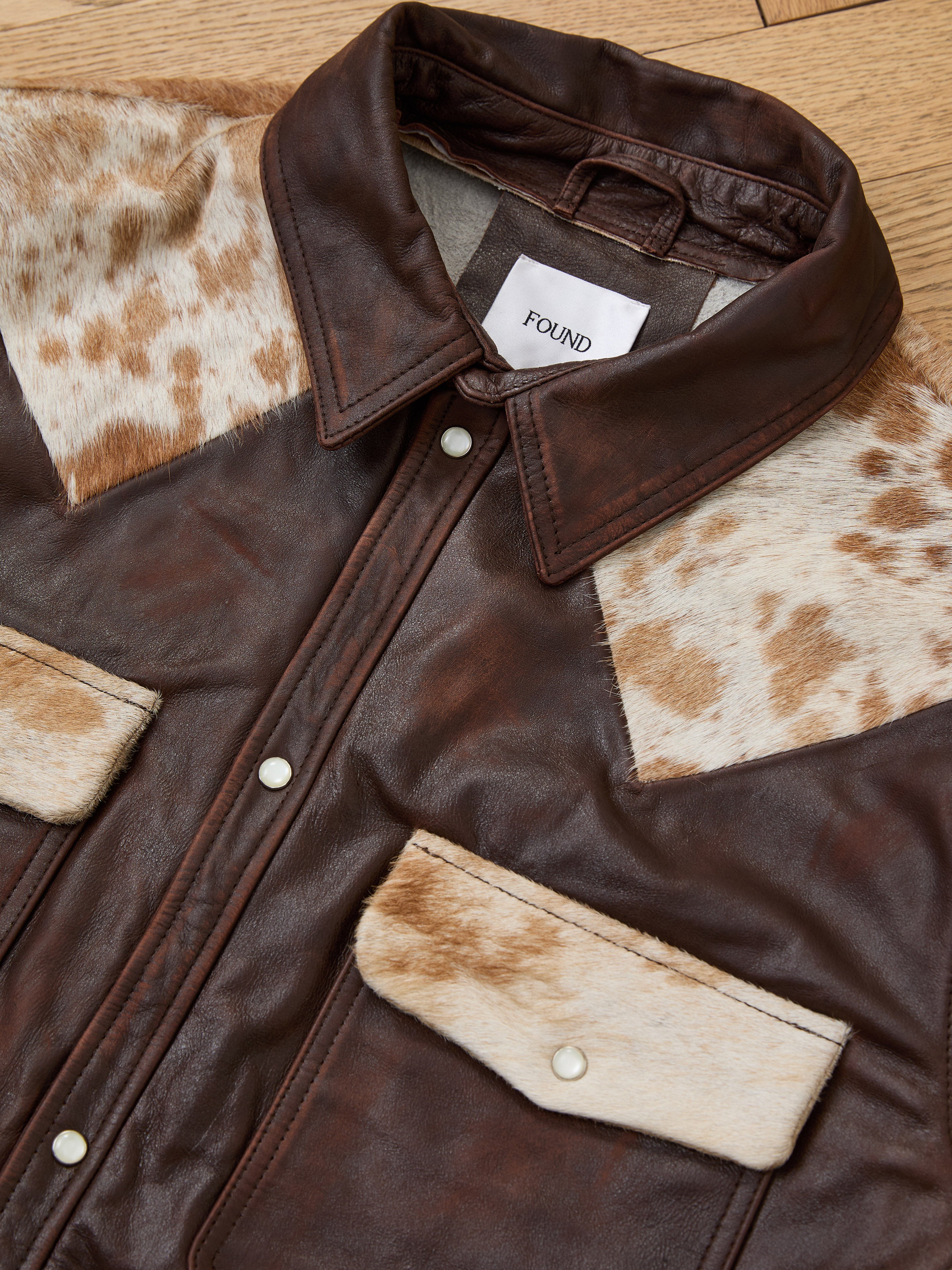 The FOUND Distressed Leather Cowhide Contrast Western Shirt, a unisex piece, features cowhide-pattern shoulder and pocket accents, silver snap buttons, and is shown laid flat on a wooden surface.