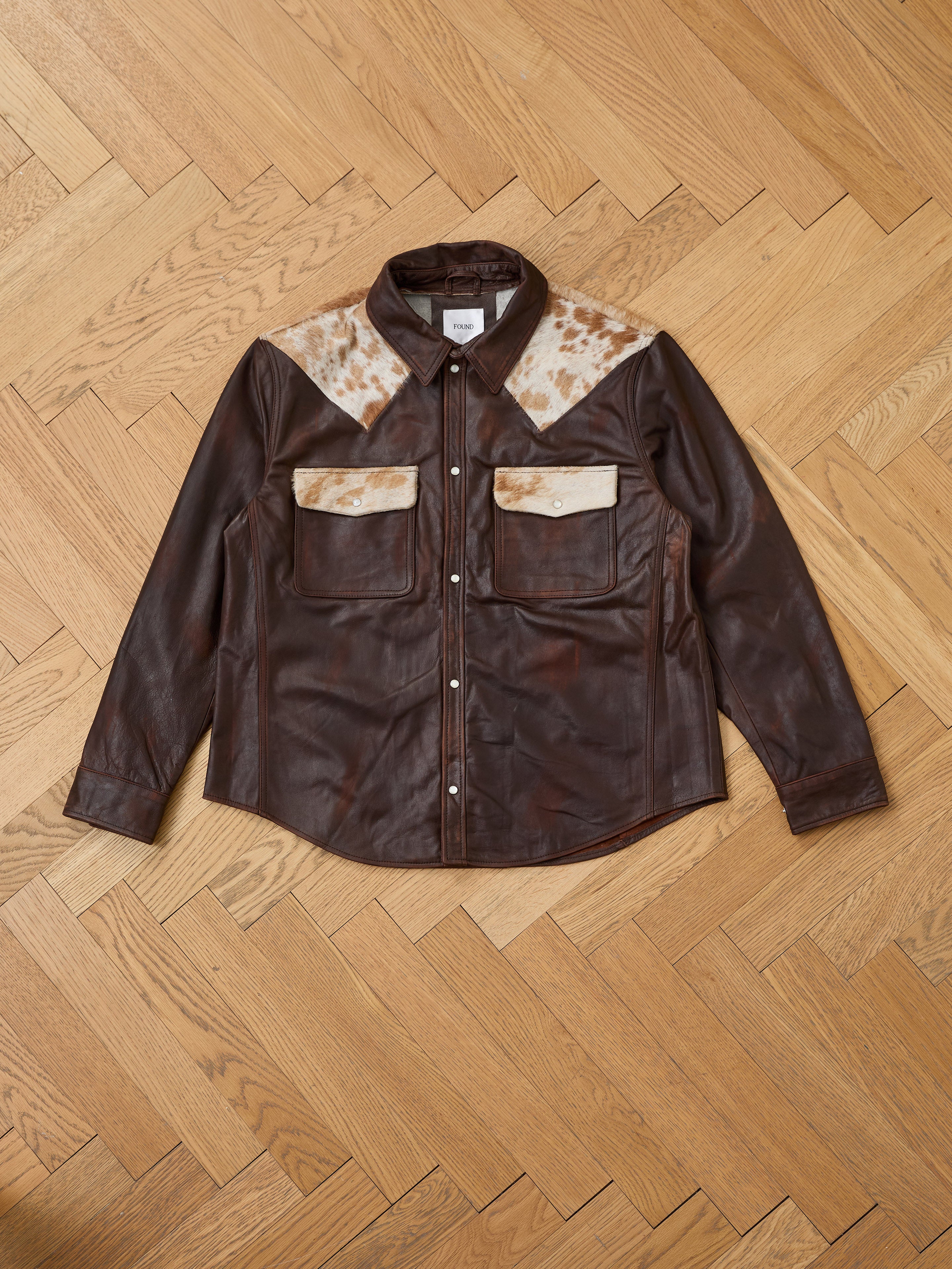 The FOUND Distressed Leather Cowhide Contrast Western Shirt features brown leather with tan and white accents—a stylish unisex western shirt shown on a herringbone wooden floor, perfect for any wardrobe.