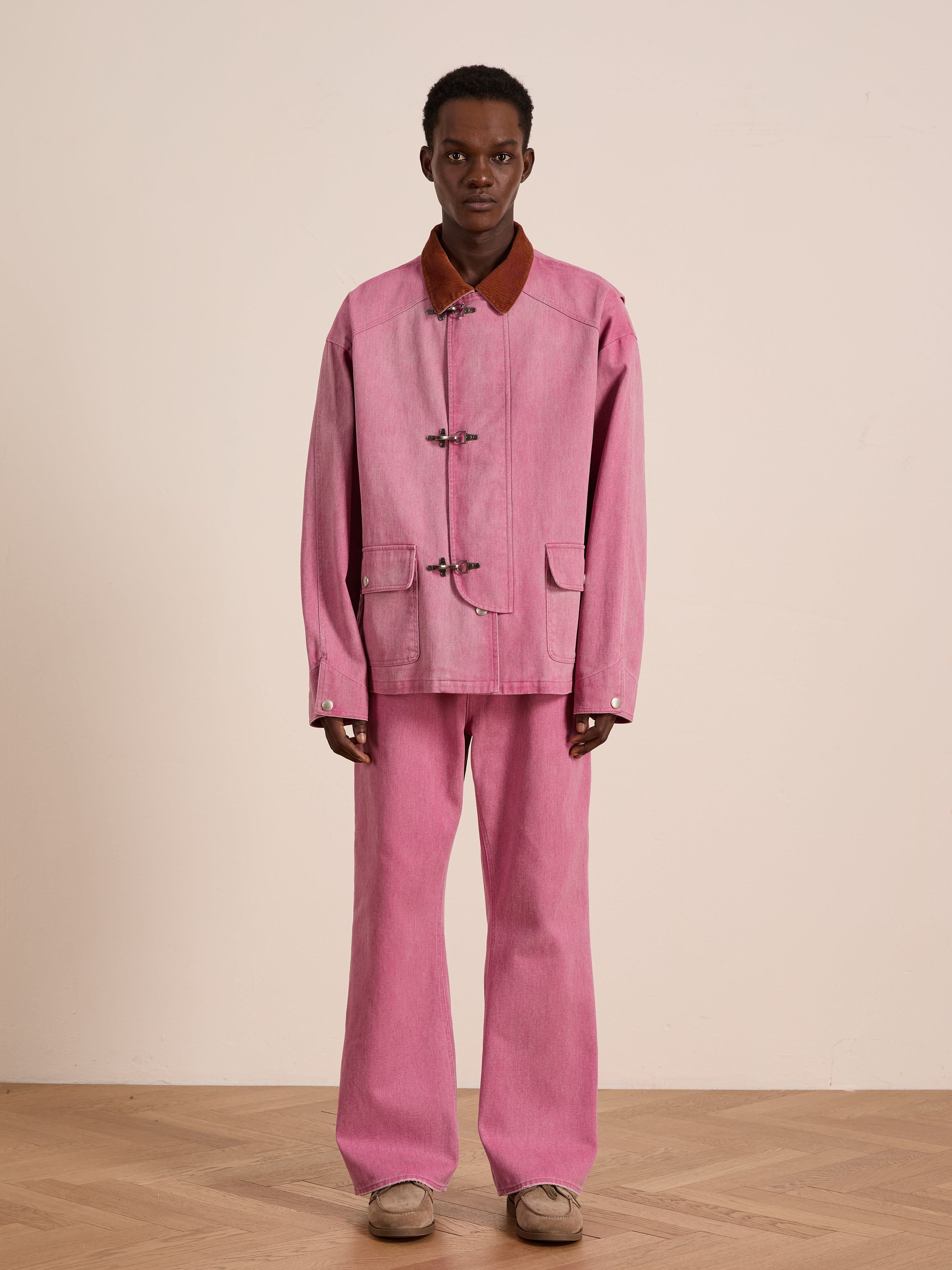 Someone stands against a plain wall wearing the Found Washed Fireman Clasp Workshop Jacket with matching wide-leg pink pants and beige shoes.
