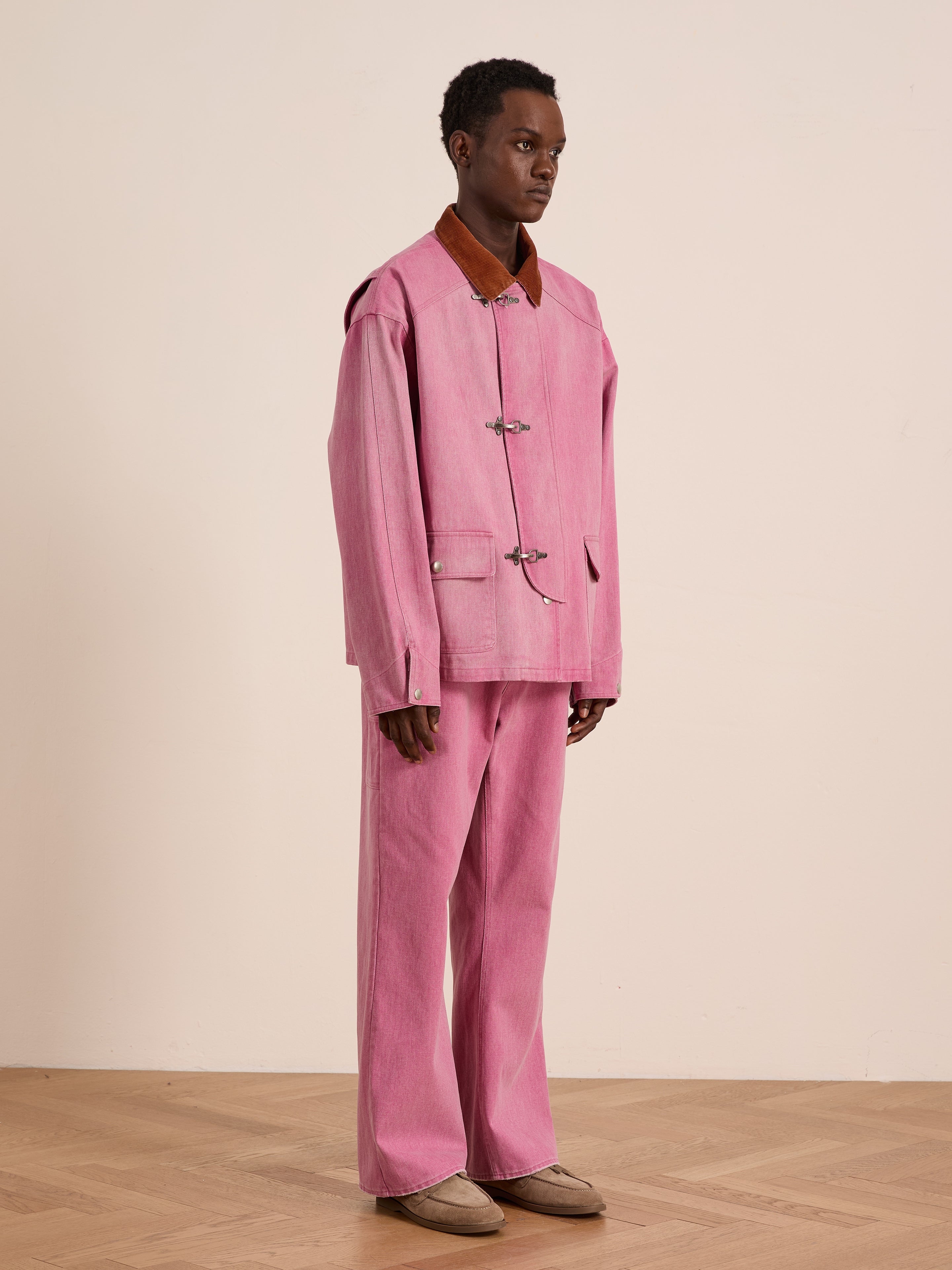 A person stands indoors against a plain background wearing the Found Washed Fireman Clasp Workshop Jacket—loose-fitting with a pink corduroy collar—paired with matching wide-leg pink pants and beige shoes.