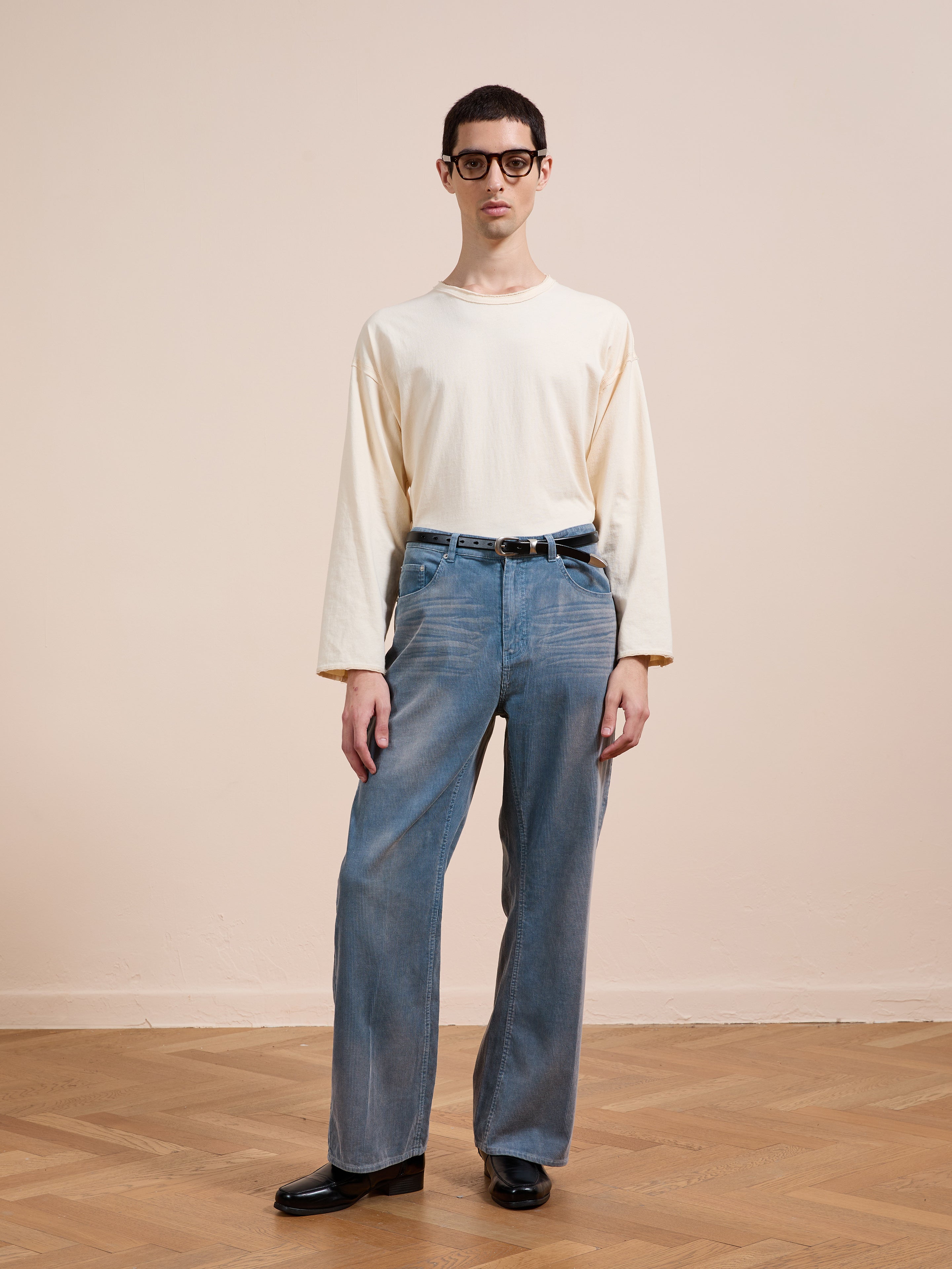 A person with short dark hair and glasses stands indoors on a wooden floor, wearing a cream long-sleeve shirt and FOUND Corduroy Flare Work Pants in vintage blue, with black shoes, against a beige wall background.