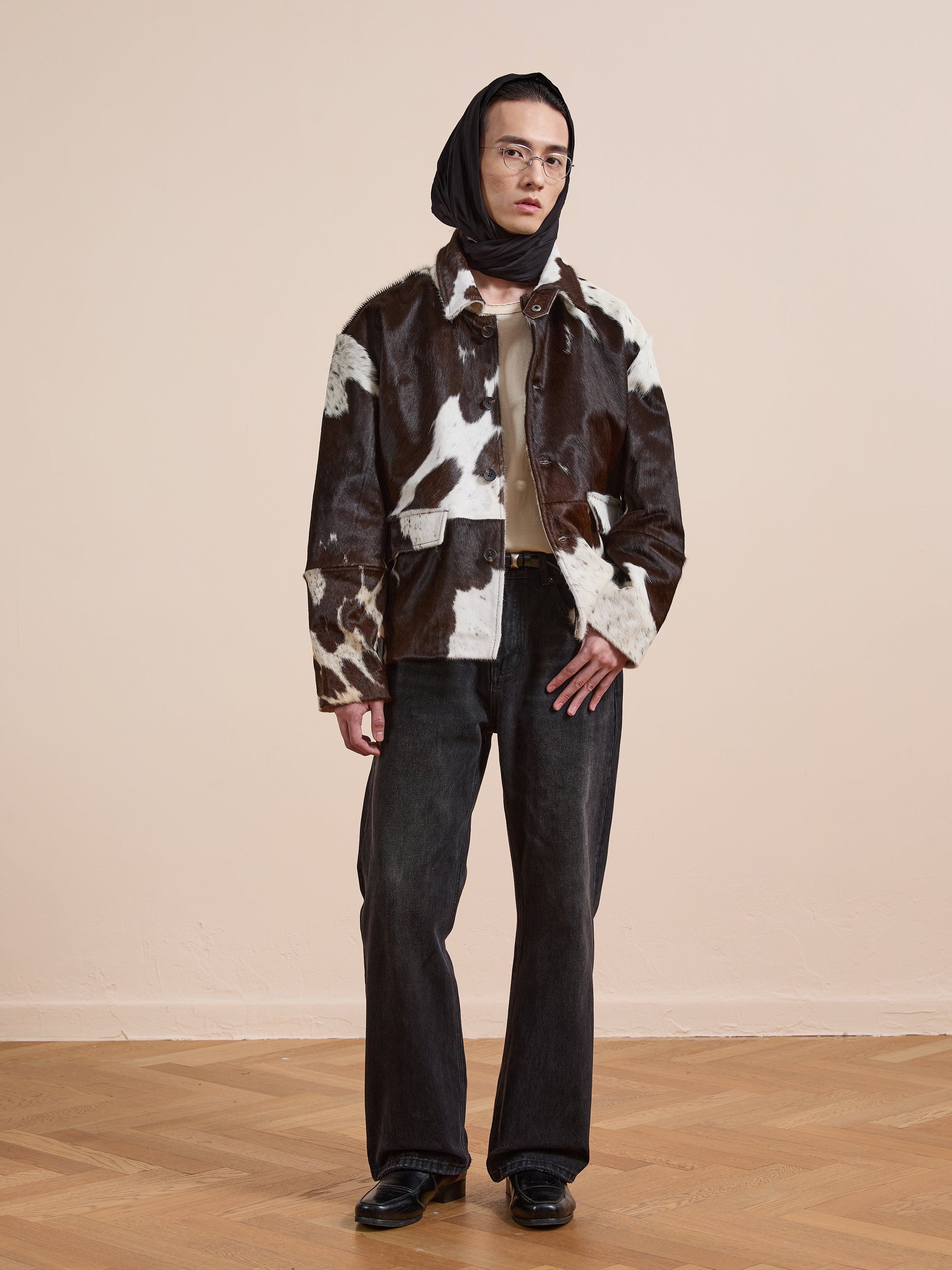 Person indoors wearing the FOUND Cowhide Prairie Utility Jacket, dark jeans, beige top, black headscarf, glasses, and black shoes on a wooden floor against a plain light wall.
