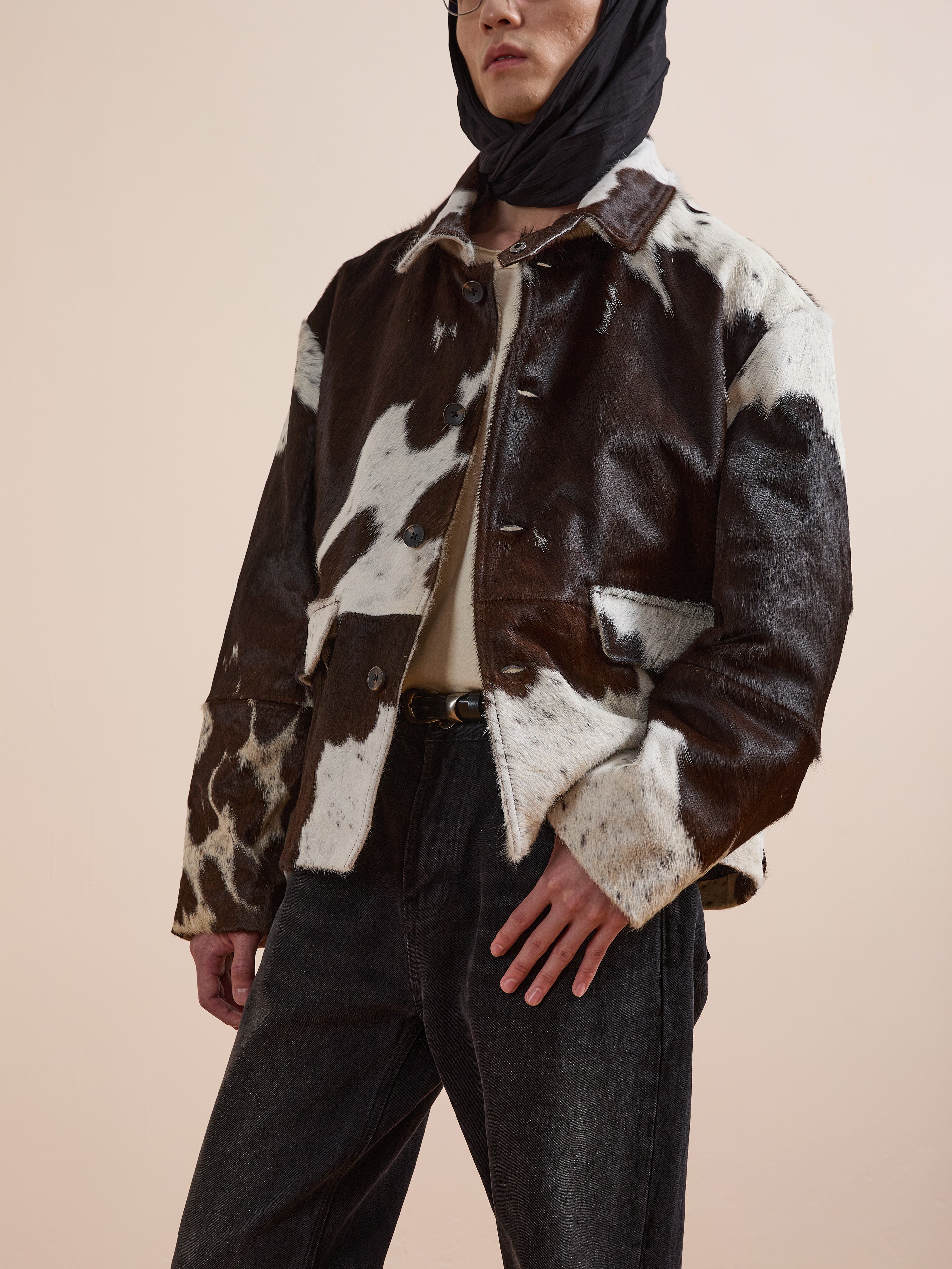 A person in the FOUND Cowhide Prairie Utility Jacket, black jeans, beige shirt, and a black headscarf poses against a plain beige background. The distinctive leather jacket brings standout style to the minimalist scene.