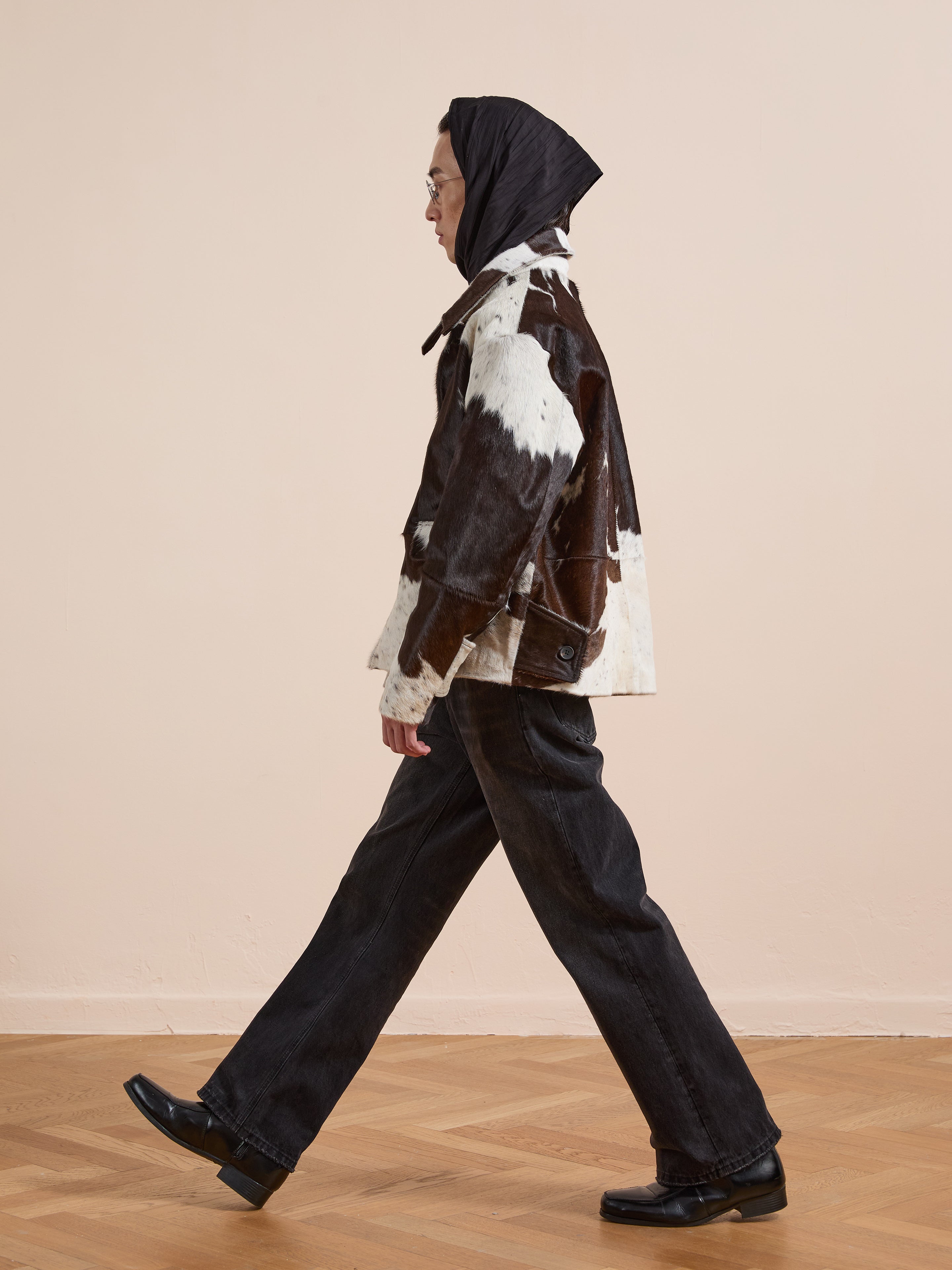 A person wears the FOUND Cowhide Prairie Utility Jacket in black and white, styled with black pants, shoes, and a headscarf, walking indoors on a wooden floor against a beige wall.