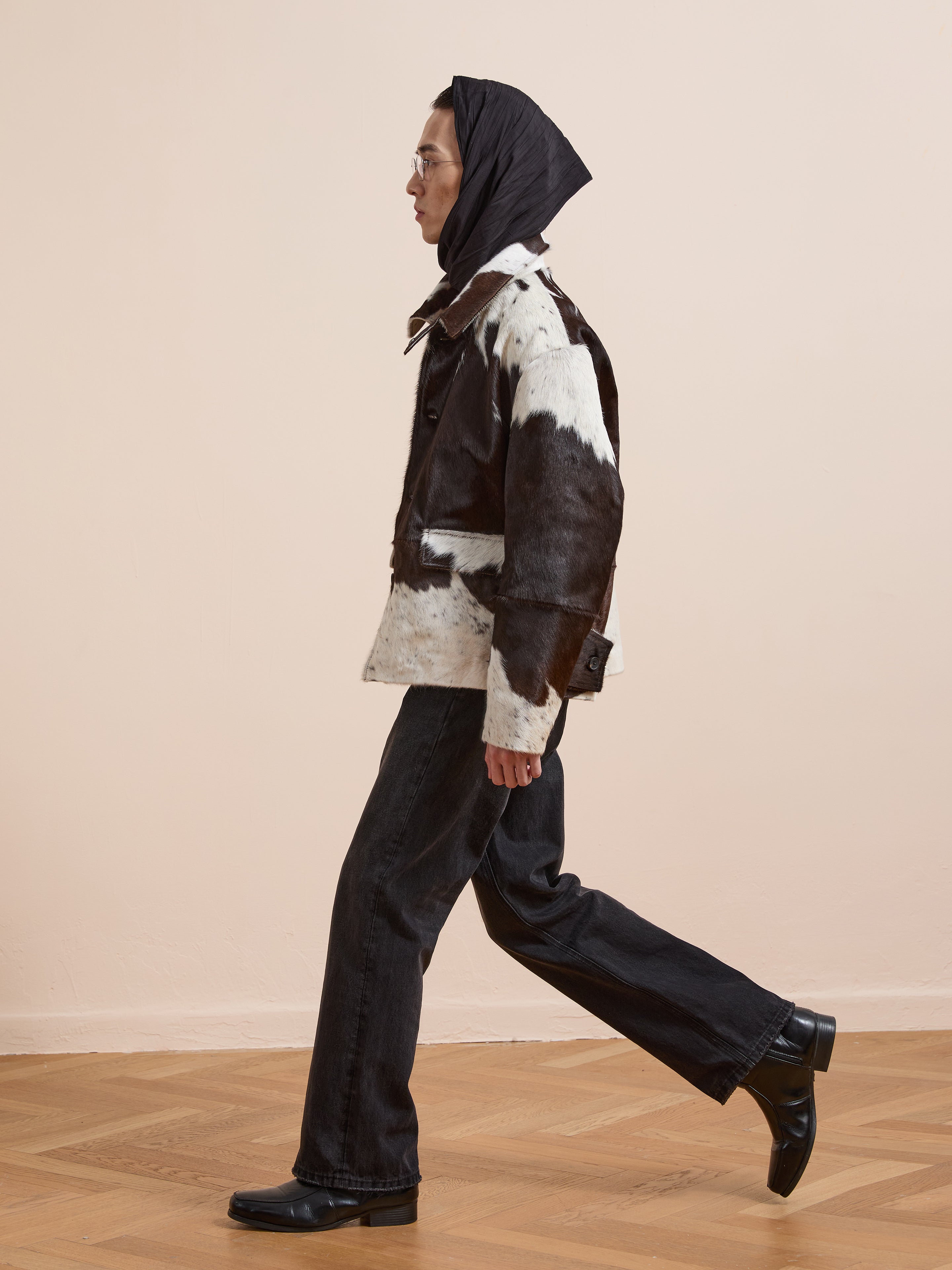 A person wearing a black headscarf, the FOUND Cowhide Prairie Utility Jacket, black jeans, and black shoes walks indoors on a wooden floor with a plain light wall in the background.