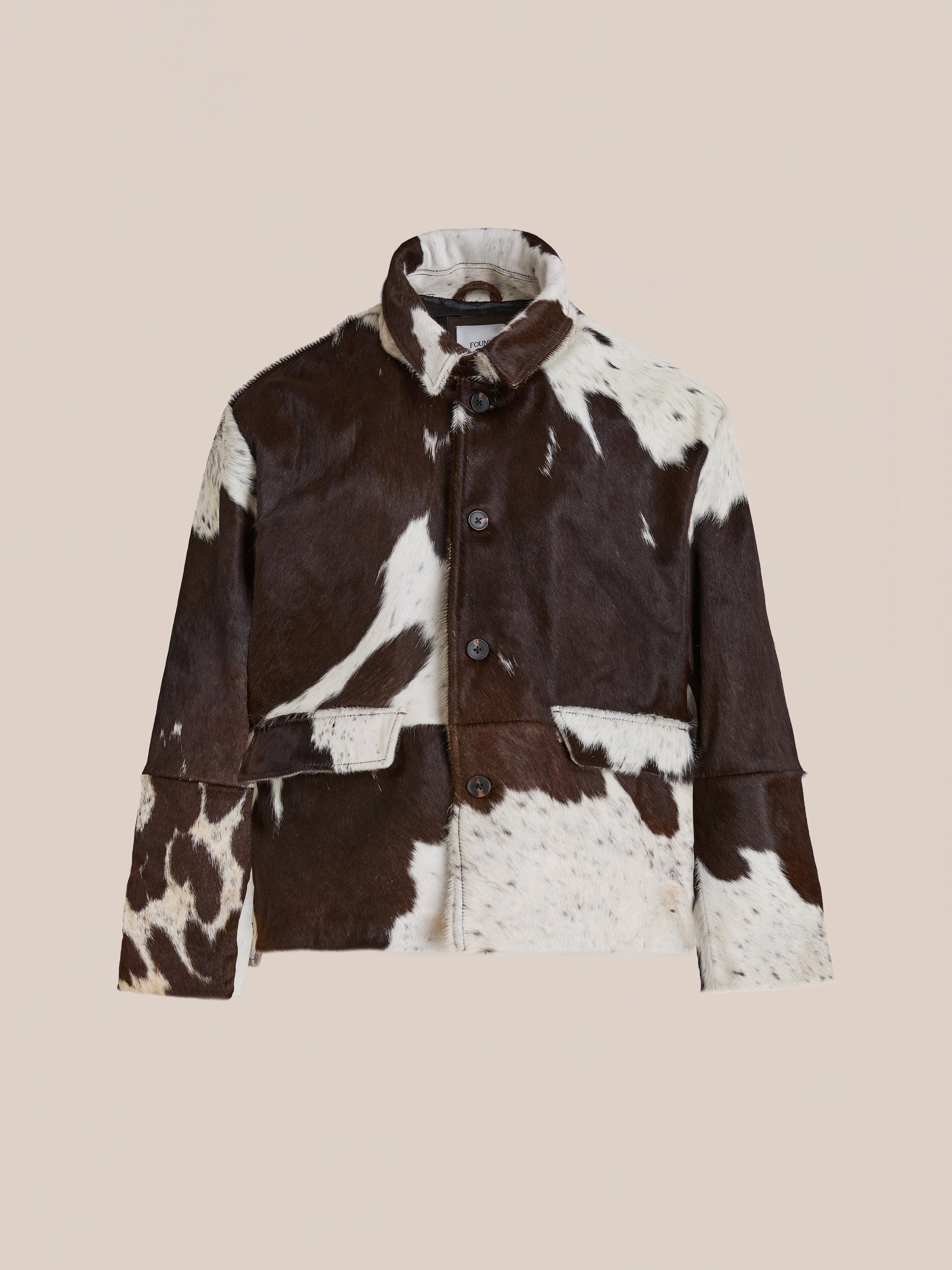 The FOUND Cowhide Prairie Utility Jacket, featuring a button front and collar in brown and white leather, is shown against a plain beige background.