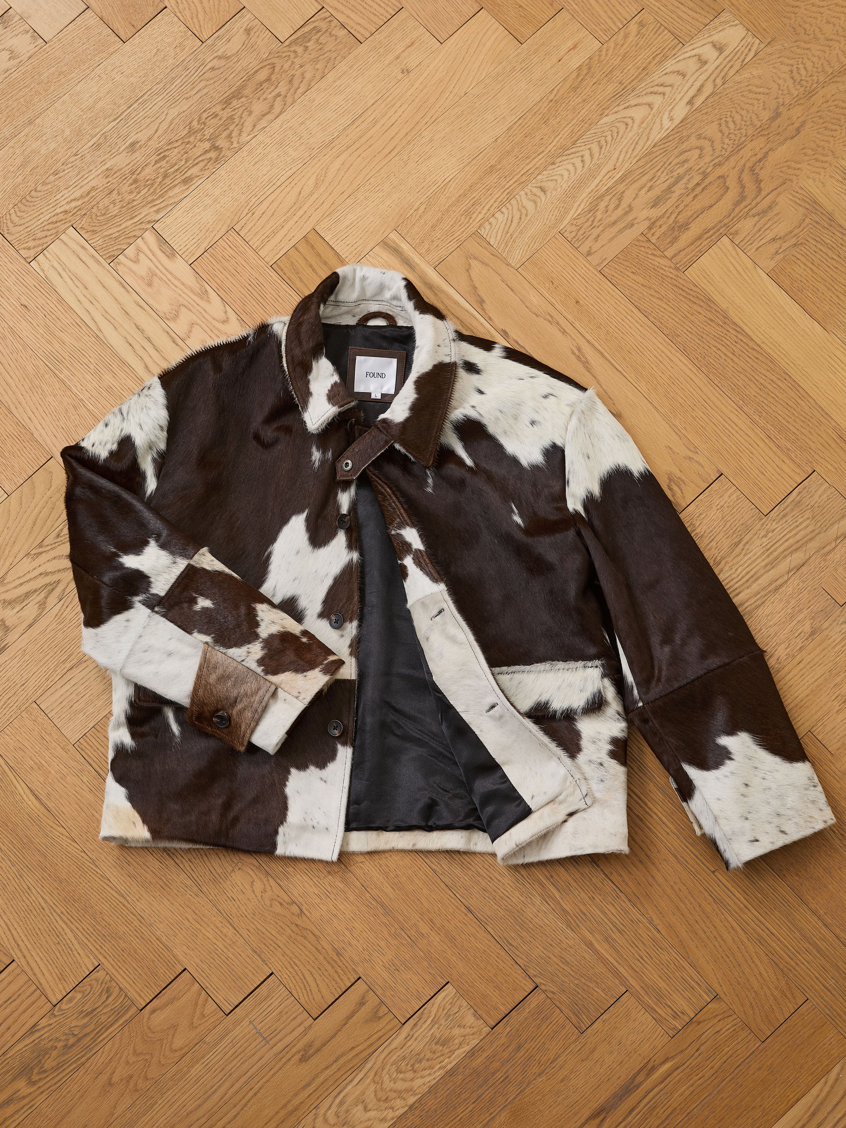 The FOUND Cowhide Prairie Utility Jacket features brown and white cowhide leather with a button-up front and collar, displayed flat on a wood herringbone floor.