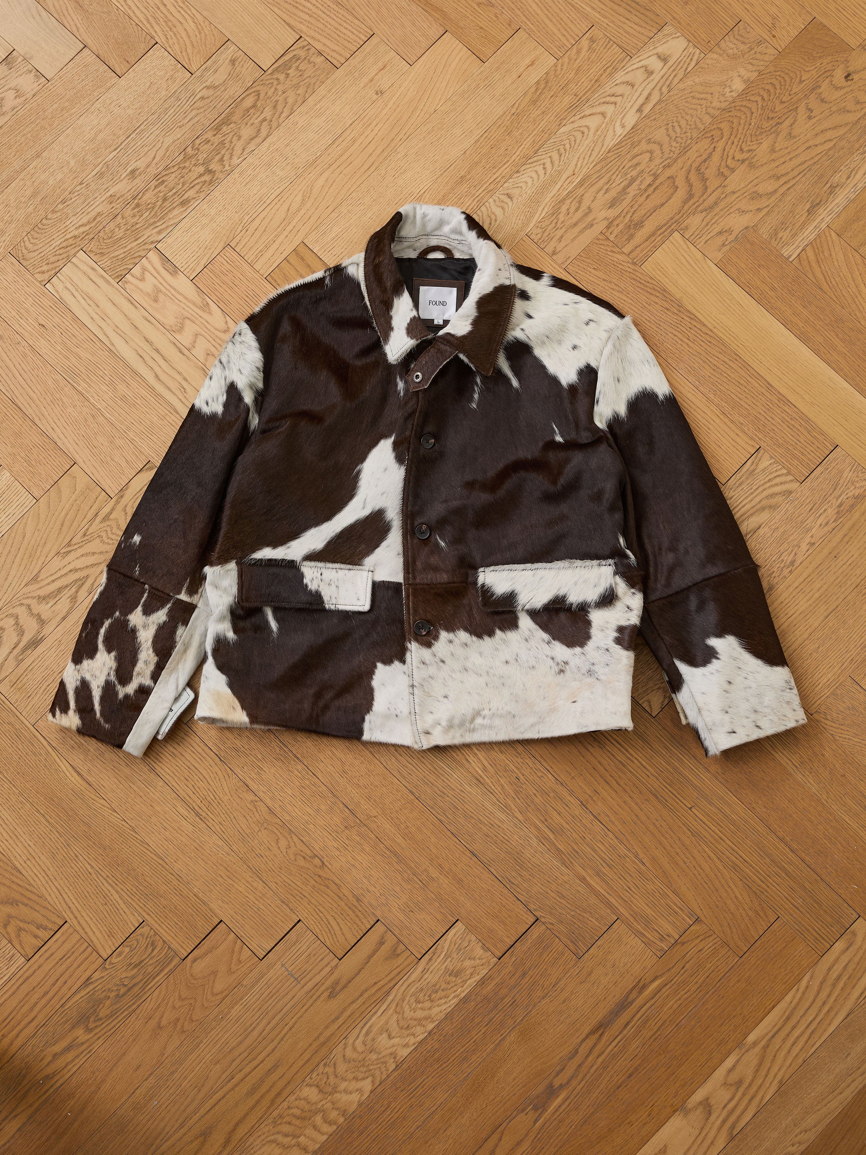 The Cowhide Prairie Utility Jacket by FOUND features a brown and white cowhide pattern, button closure, and collar. Displayed on a light wood herringbone floor, this standout leather jacket offers bold style and unique charm.