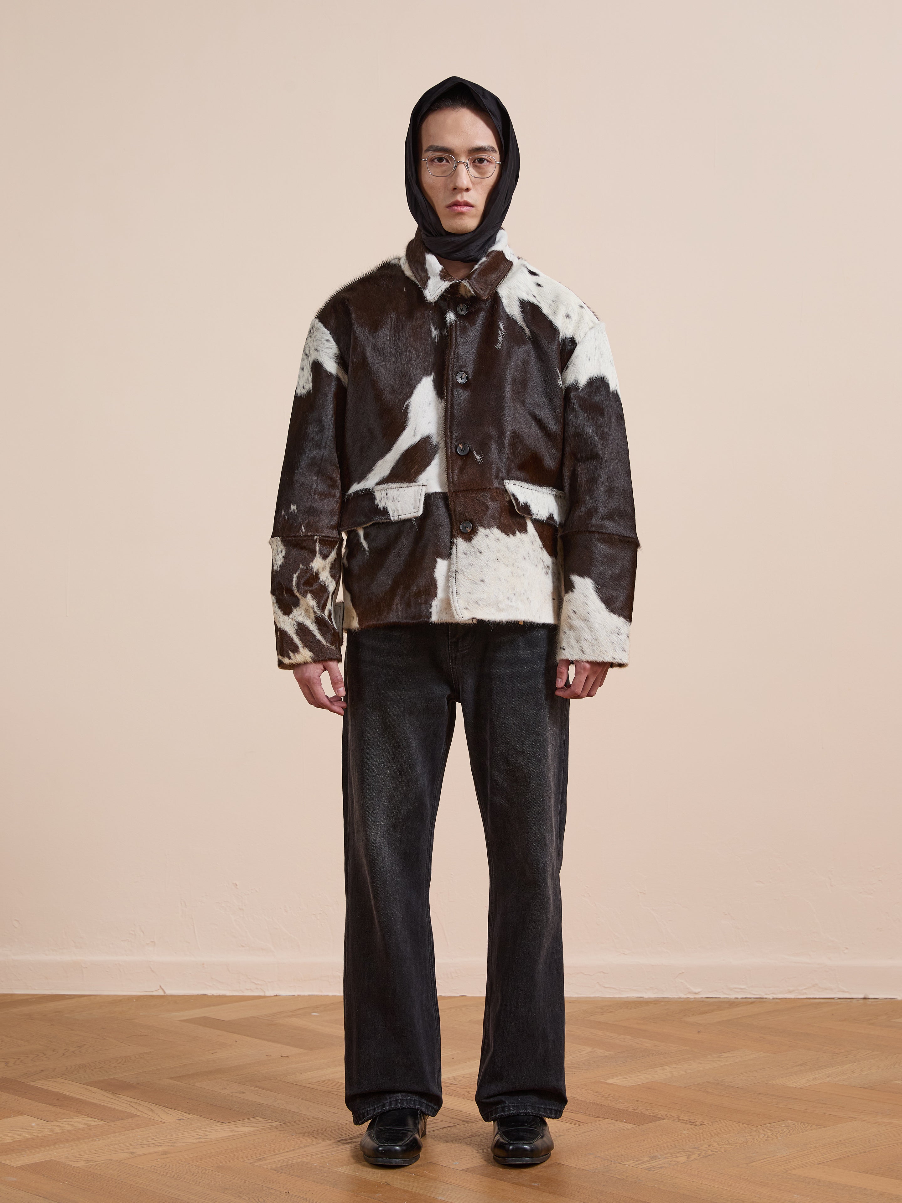 A person indoors models the FOUND Cowhide Prairie Utility Jacket, featuring a black and white cowhide print, styled with a black hood, dark jeans, and black shoes against a plain light wall and wooden floor.