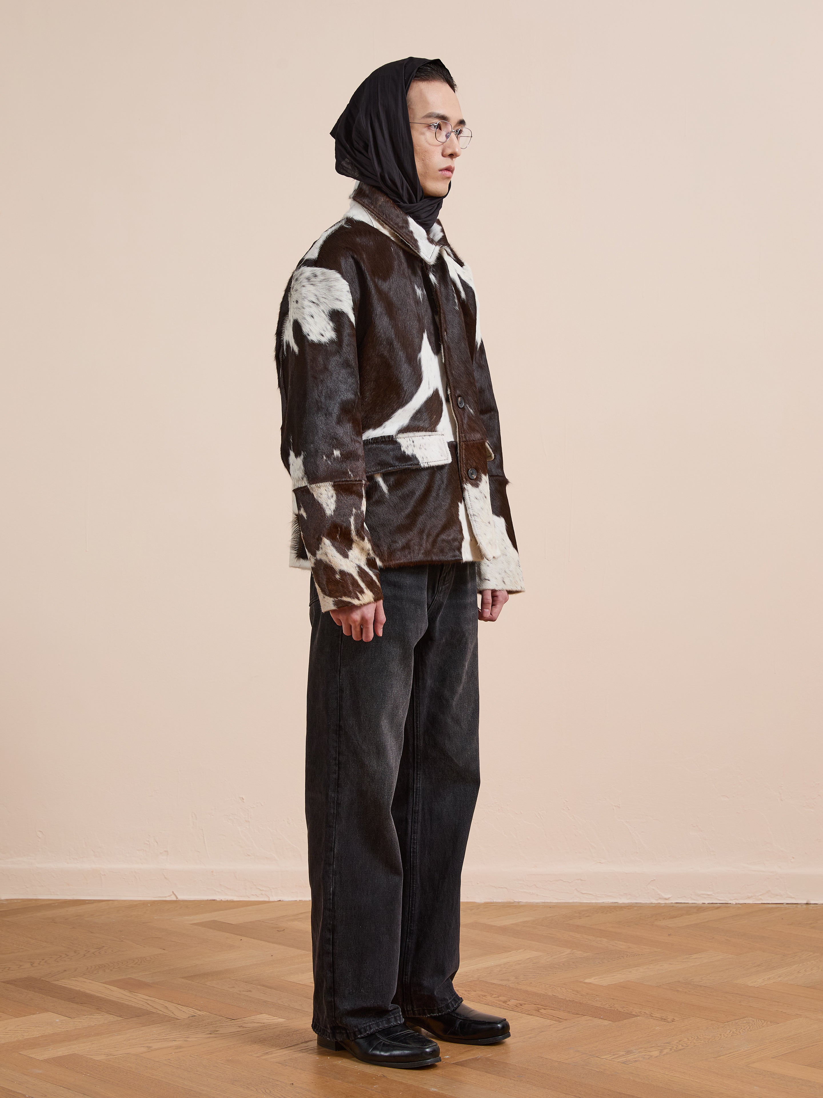 A person stands indoors against a plain beige background wearing FOUND’s Cowhide Prairie Utility Jacket, featuring a bold black and white pattern, paired with black wide-leg pants, black shoes, glasses, and a black headscarf.