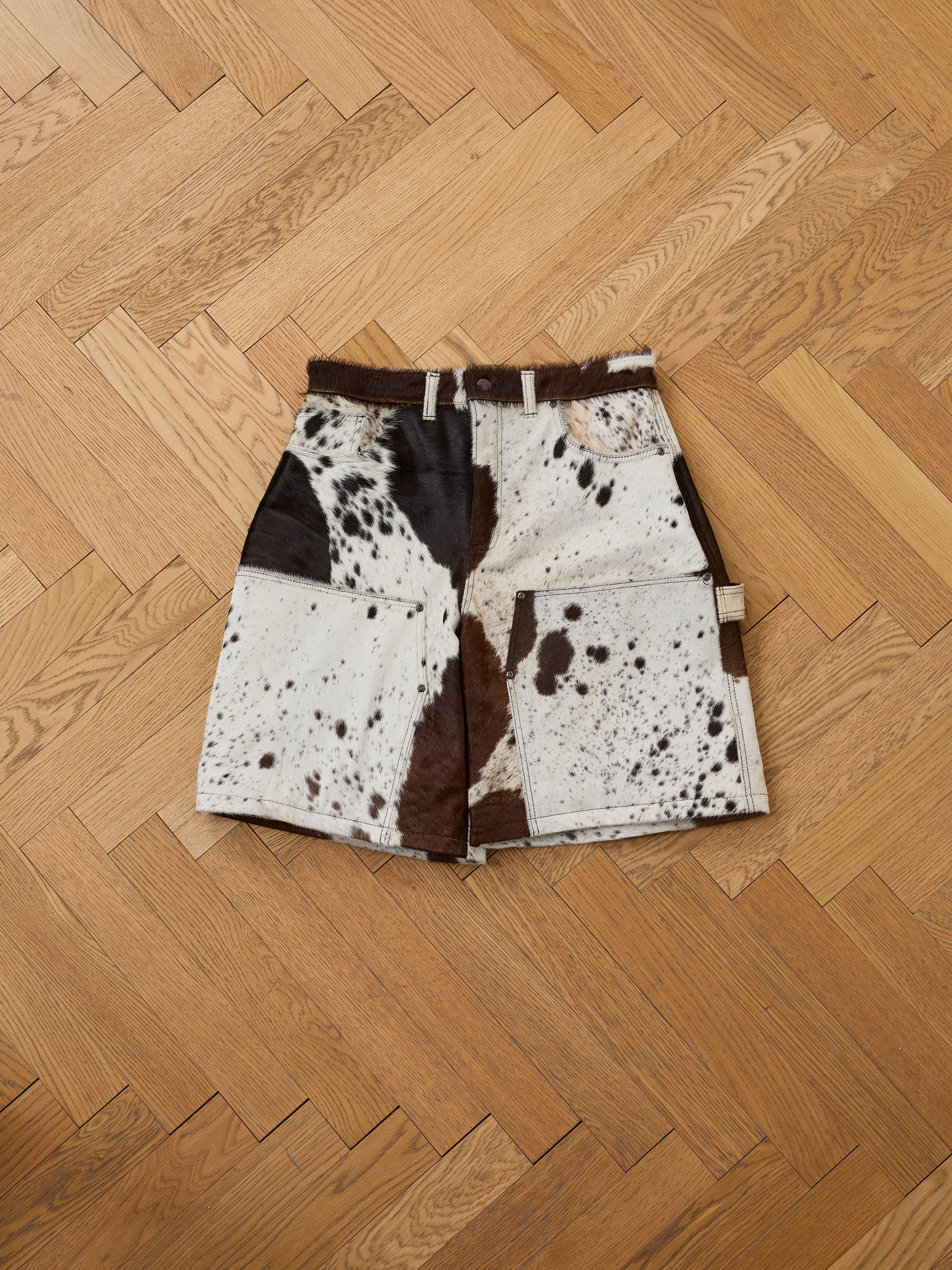 The Found Cowhide Prairie Carpenter Shorts are unisex, relaxed fit brown and white shorts with large front pockets, shown on a herringbone wood floor.