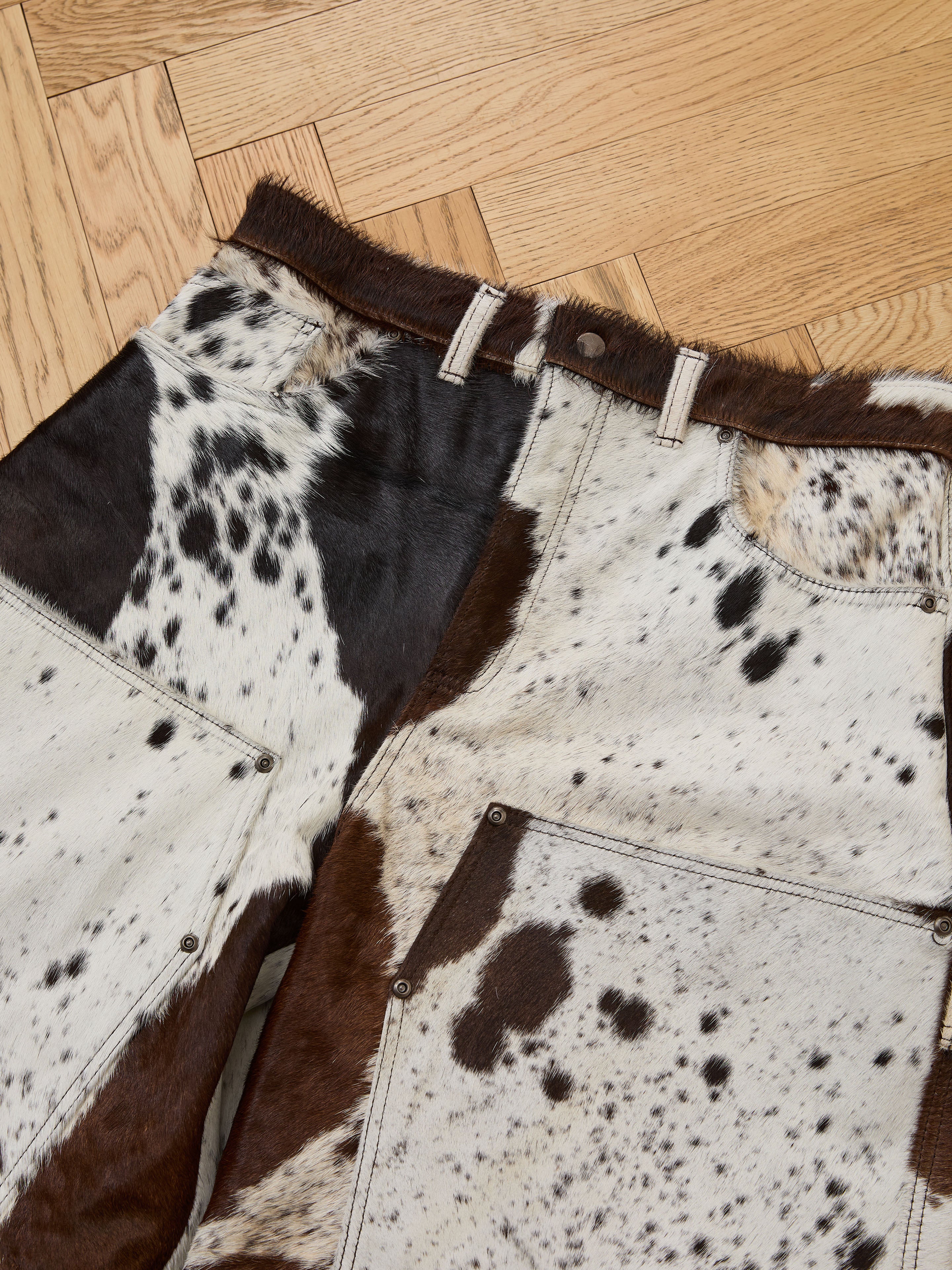 The Found Cowhide Prairie Carpenter Shorts are unisex, relaxed fit shorts made from genuine brown and white spotted cowhide leather, shown laid flat on a wooden parquet floor.