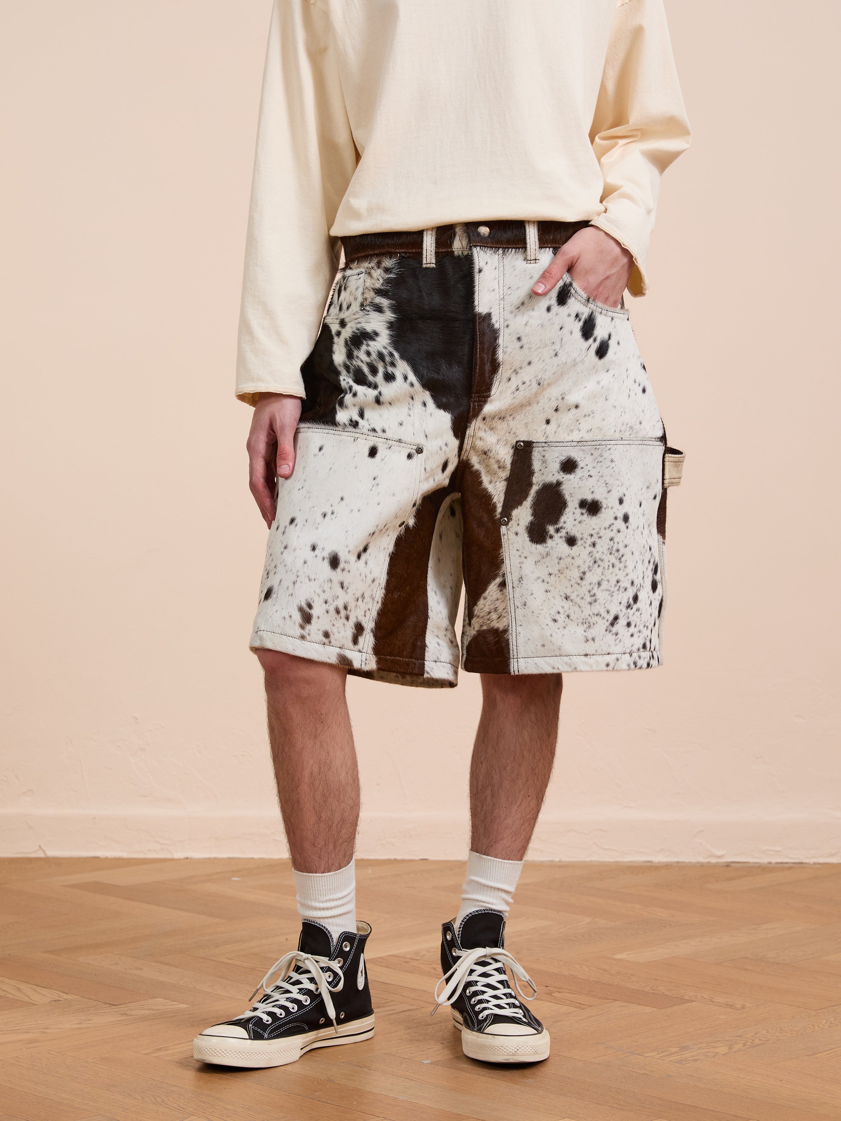 A person in a cream long-sleeve shirt, Found Cowhide Prairie Carpenter Shorts, white socks, and black high-top sneakers stands on a wooden floor against a beige wall.