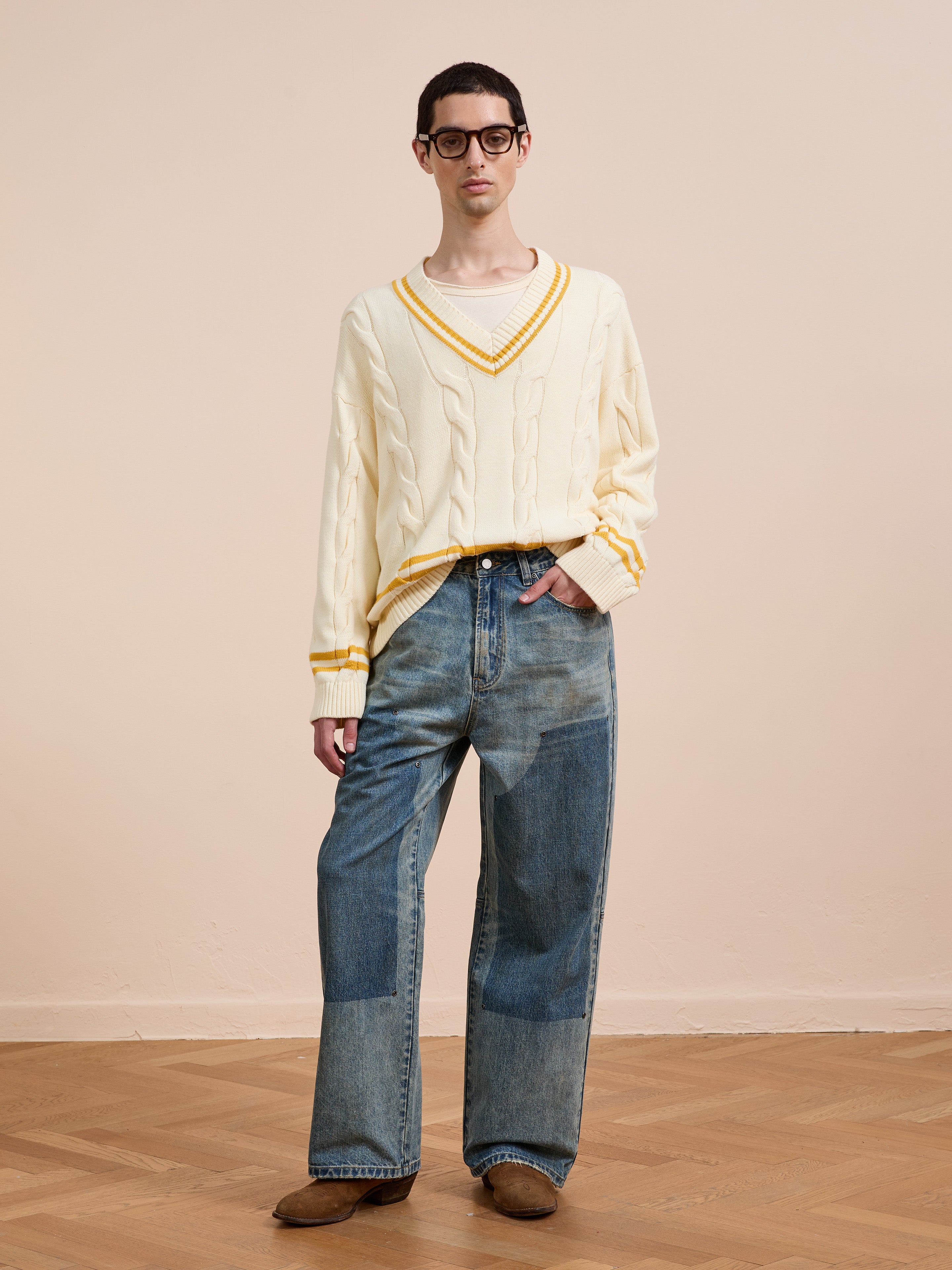 A person stands indoors on a wooden floor against a beige wall, wearing the FOUND V-Neck Cricket Sweater in cream with yellow trim, paired with loose patchwork jeans, brown shoes, and black glasses.