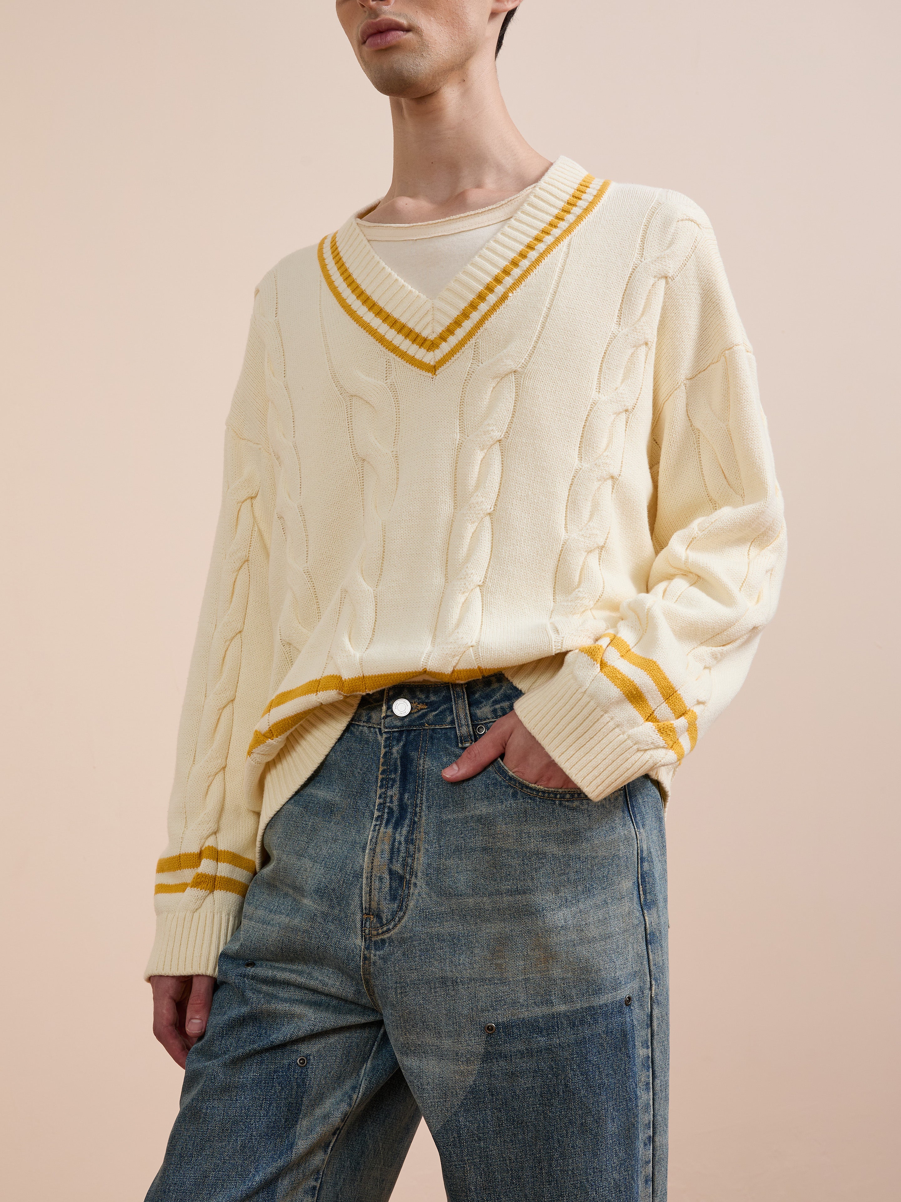 A person wears the FOUND V-Neck Cricket Sweater, featuring cream color and yellow stripes, over a beige shirt with blue jeans, standing against a neutral background.