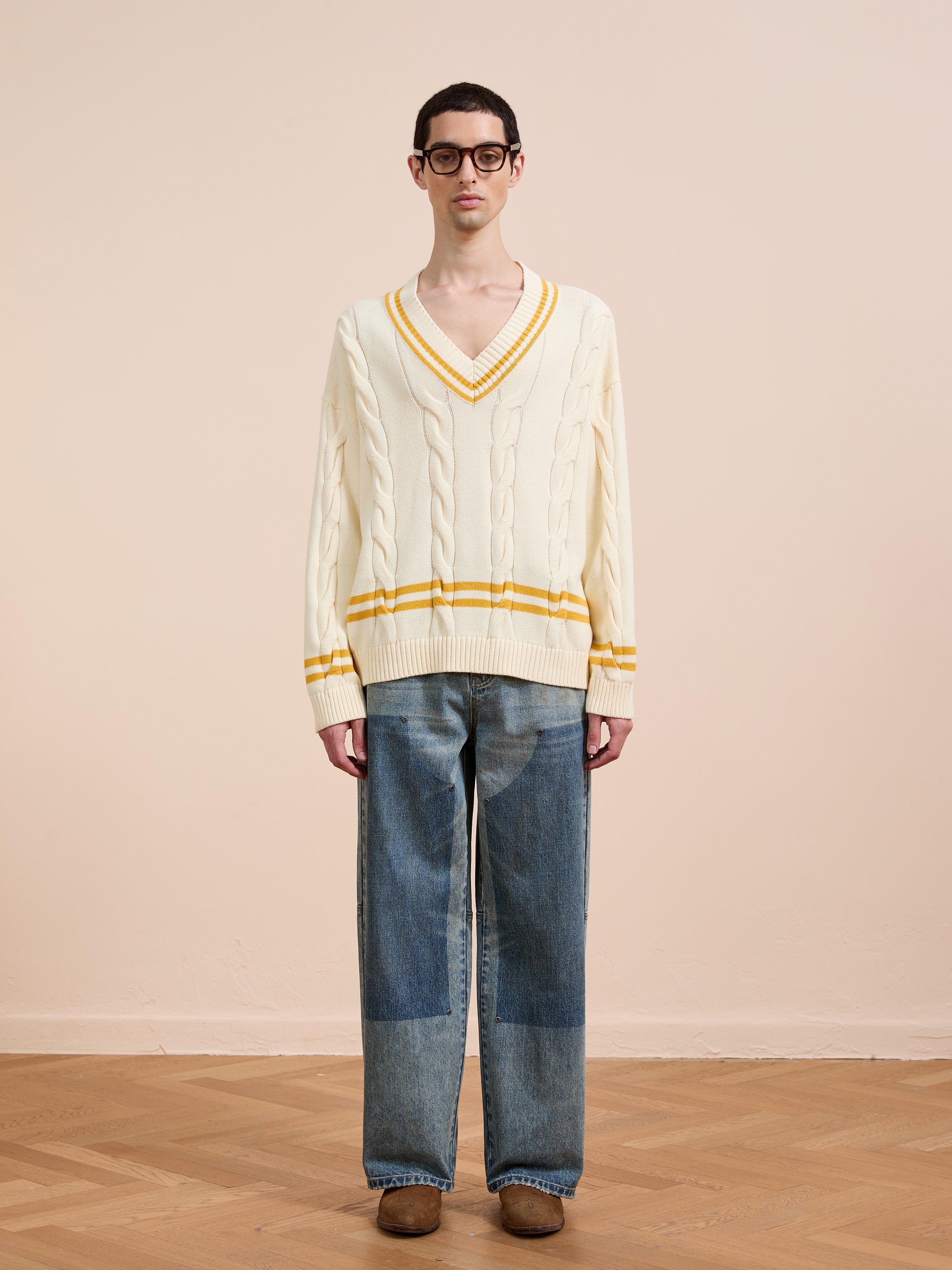 A person stands indoors against a beige background, wearing FOUND’s V-Neck Cricket Sweater in cream with yellow trim, loose blue jeans with knee patches, brown shoes, and black glasses.