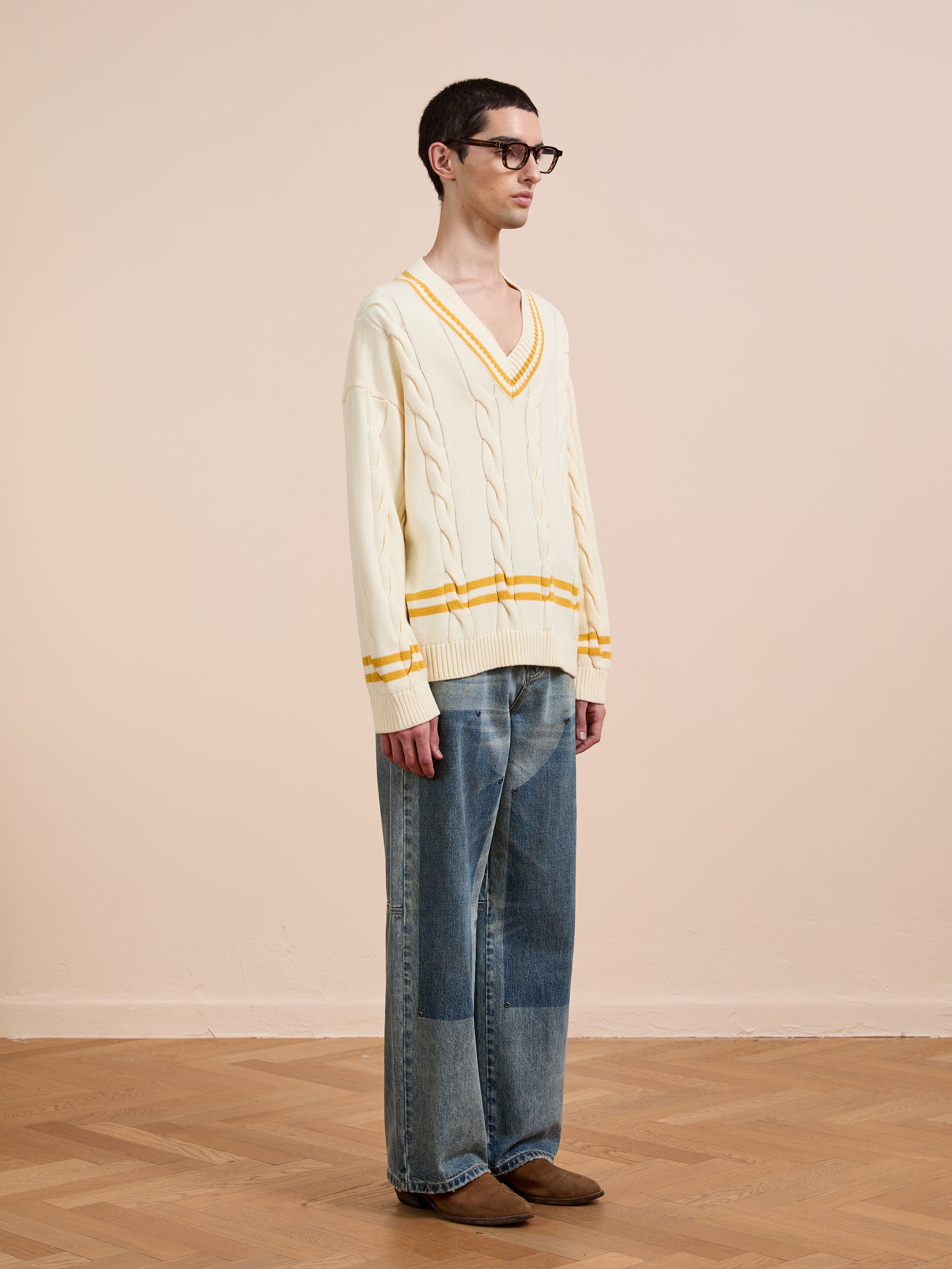 A person wearing glasses, a FOUND V-Neck Cricket Sweater in cream with yellow stripes, loose blue jeans, and brown shoes stands on a wooden floor against a plain beige wall.