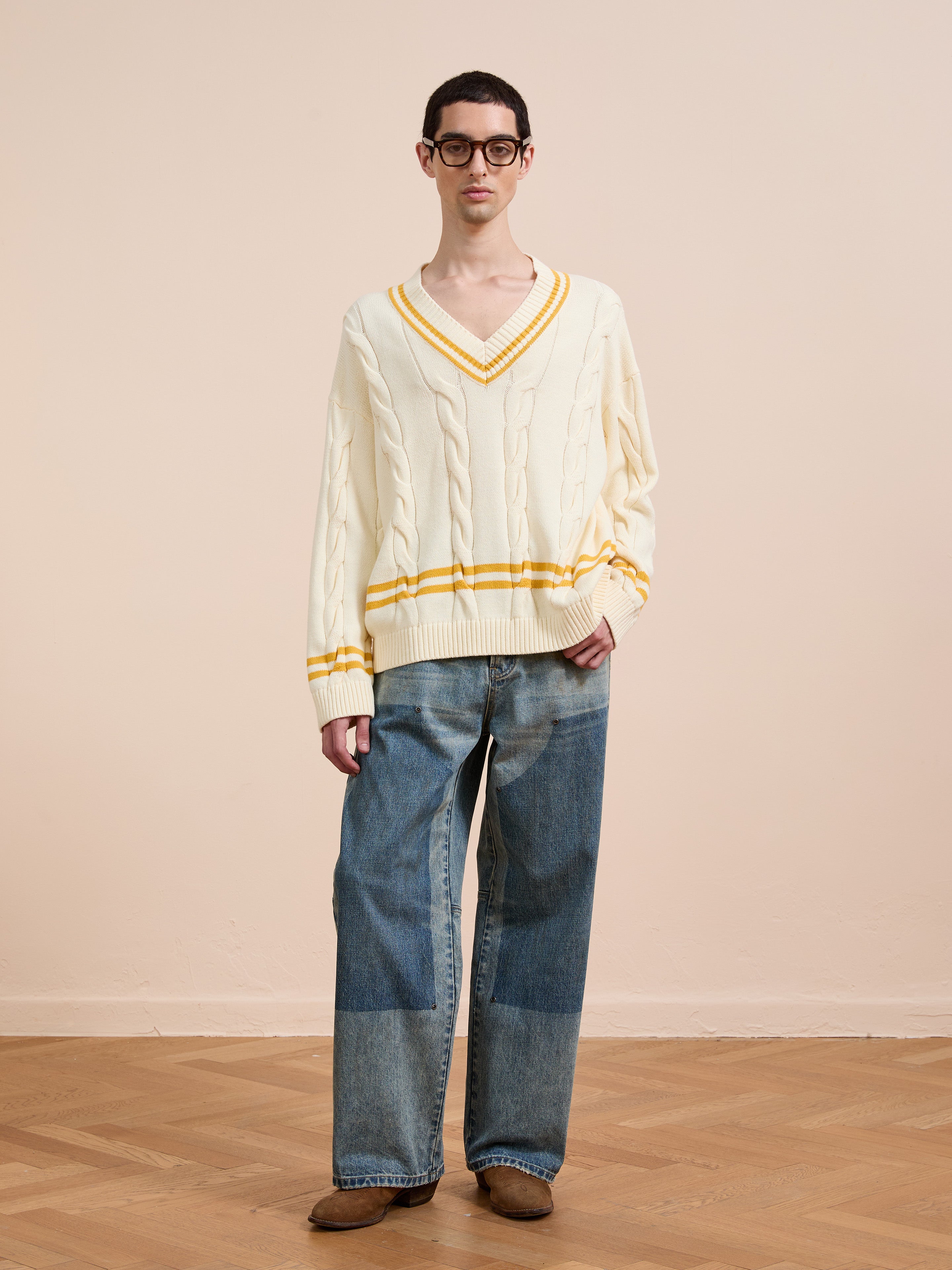 A person in the FOUND V-Neck Cricket Sweater (cream with yellow stripes), blue jeans, brown shoes, and glasses stands on a wooden floor against a beige wall.