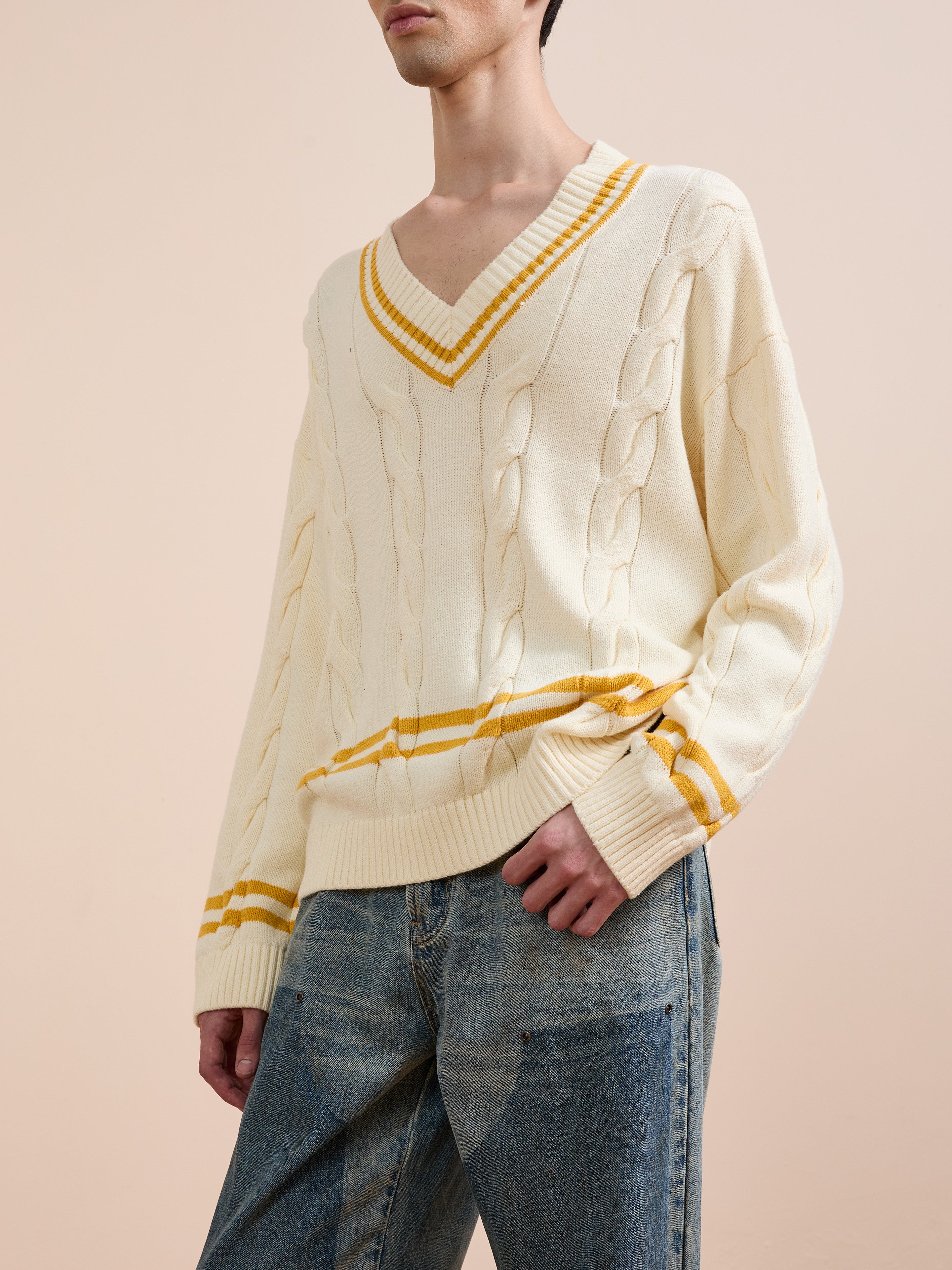 A person wears FOUND's cream-colored V-Neck Cricket Sweater with yellow stripes and blue jeans, standing against a neutral background.