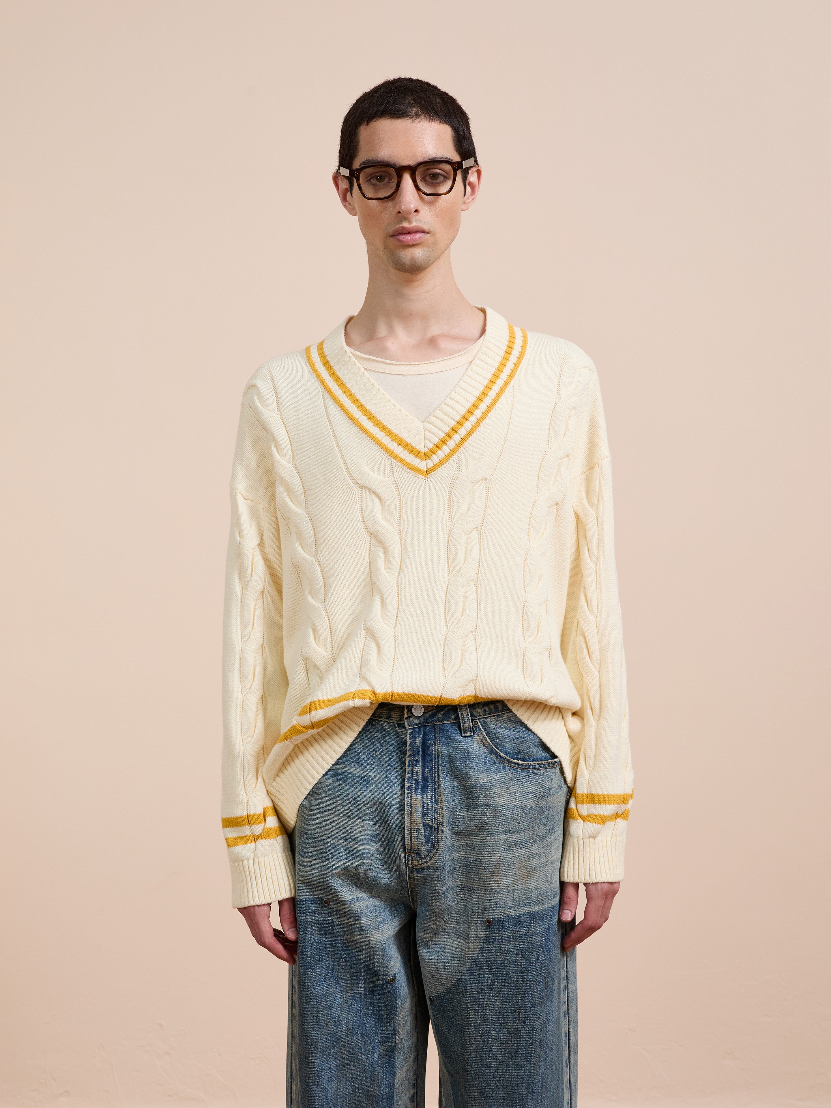 A person in glasses and blue jeans wears the FOUND V-Neck Cricket Sweater, a cream unisex cable knit with yellow trim, standing against a plain beige background.