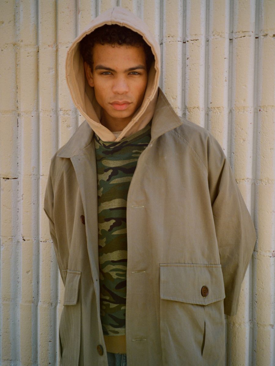 A person stands before a beige, ribbed wall, wearing the FOUND Double Layer Hoodie in tan over a camouflage shirt, vintage-style, while looking directly at the camera.