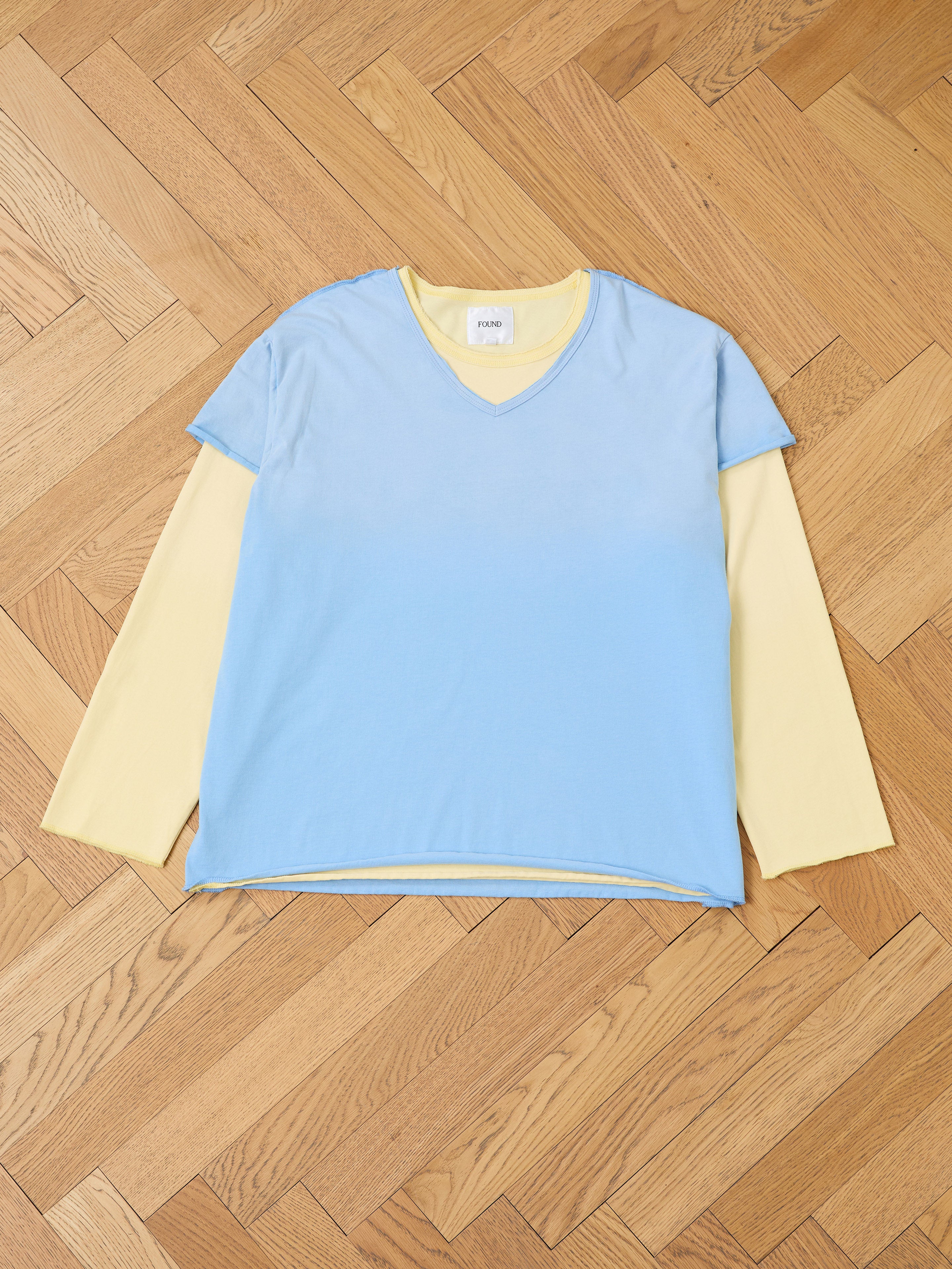 The FOUND Double Layer LS Tee features a sunfade wash unisex yellow long sleeve, layered under a short-sleeve light blue V-neck tee, showcased flat on a wooden herringbone floor.