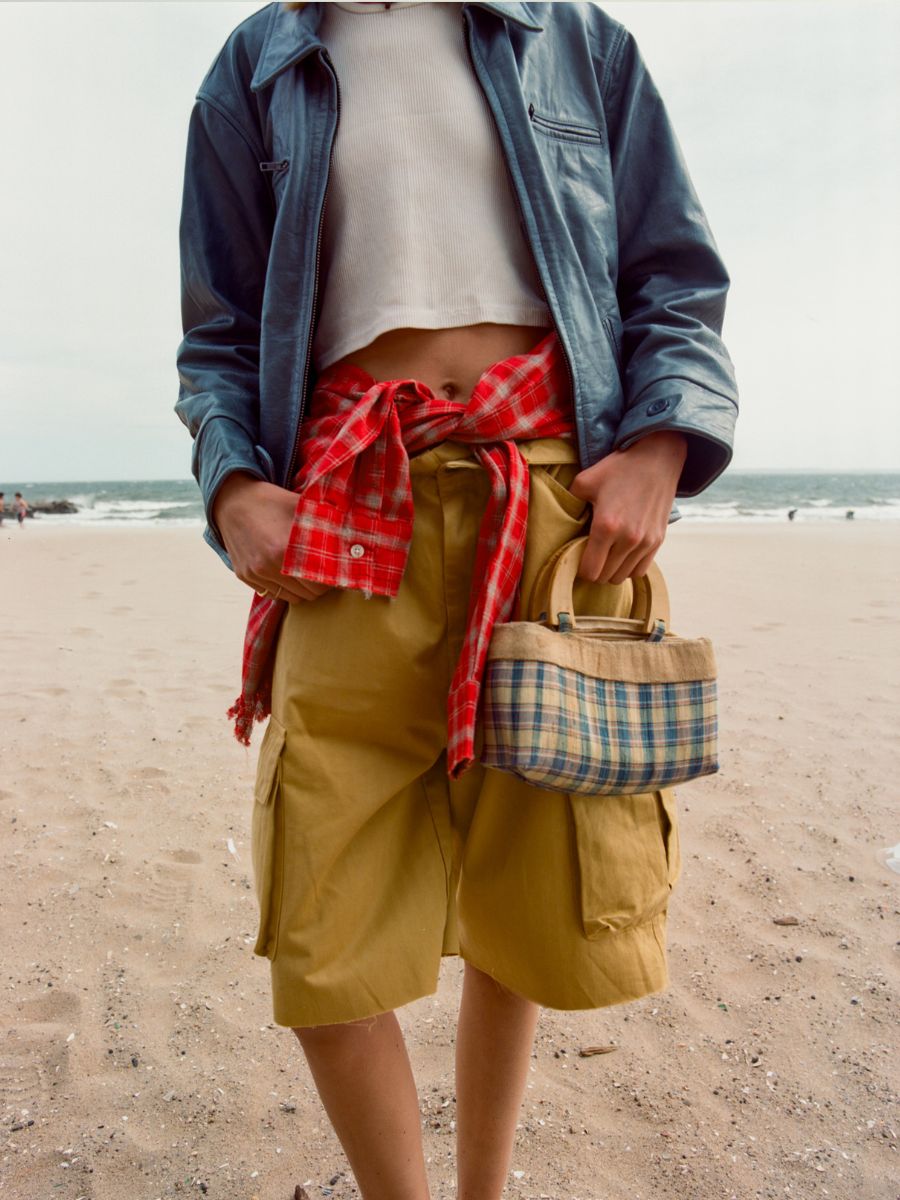 Standing on the beach in FOUND’s Marlboro Sun Faded Leather Racer Jacket, white crop top, yellow shorts, and a red plaid shirt tied at the waist, she holds a small plaid handbag—blending bold style with effortless confidence.