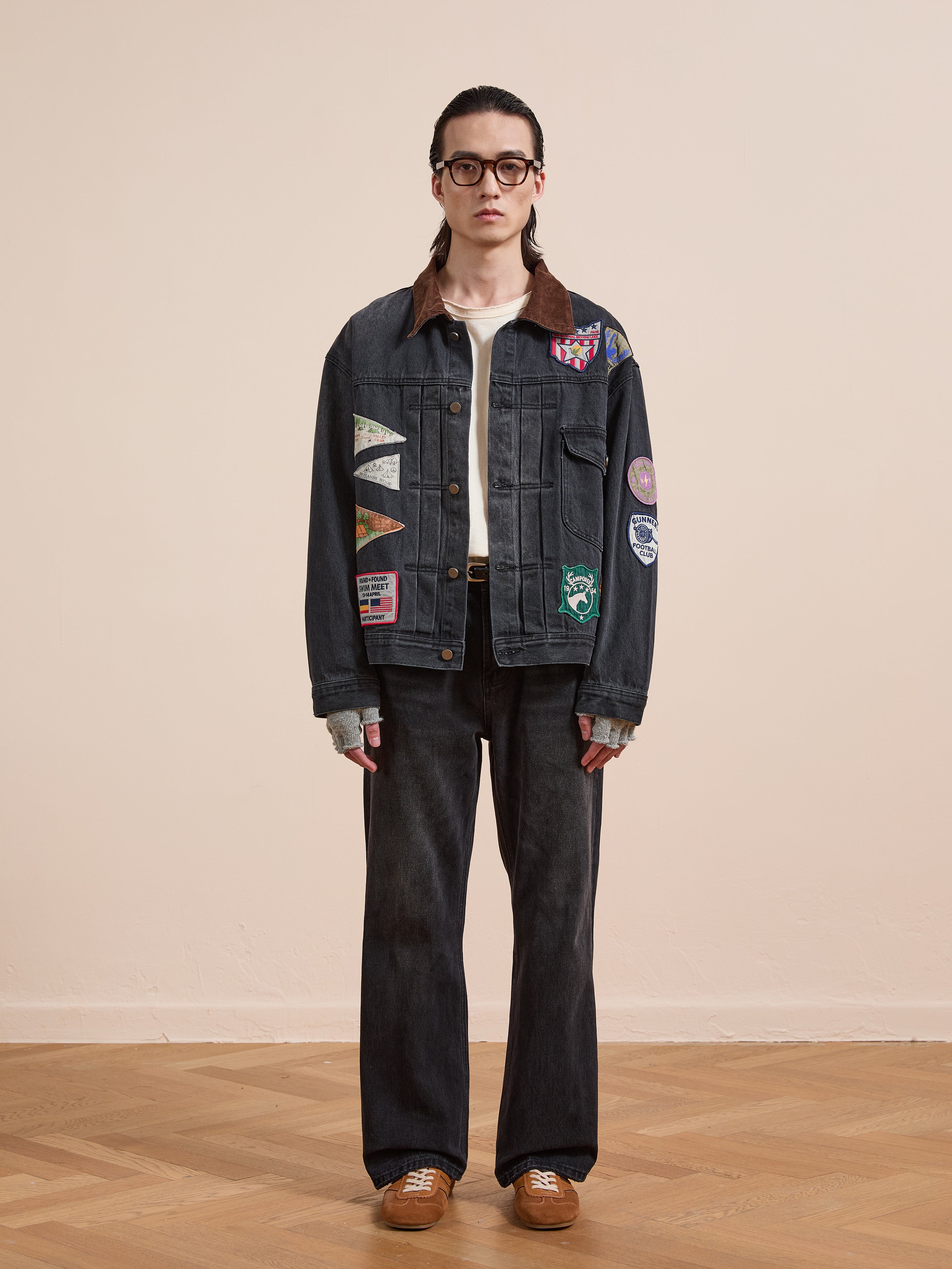 A person stands indoors against a plain background wearing the FOUND Denim Patchwork Jacket with Americana patches and a corduroy collar, paired with a white shirt, black jeans, gray fingerless gloves, brown sneakers, and glasses.