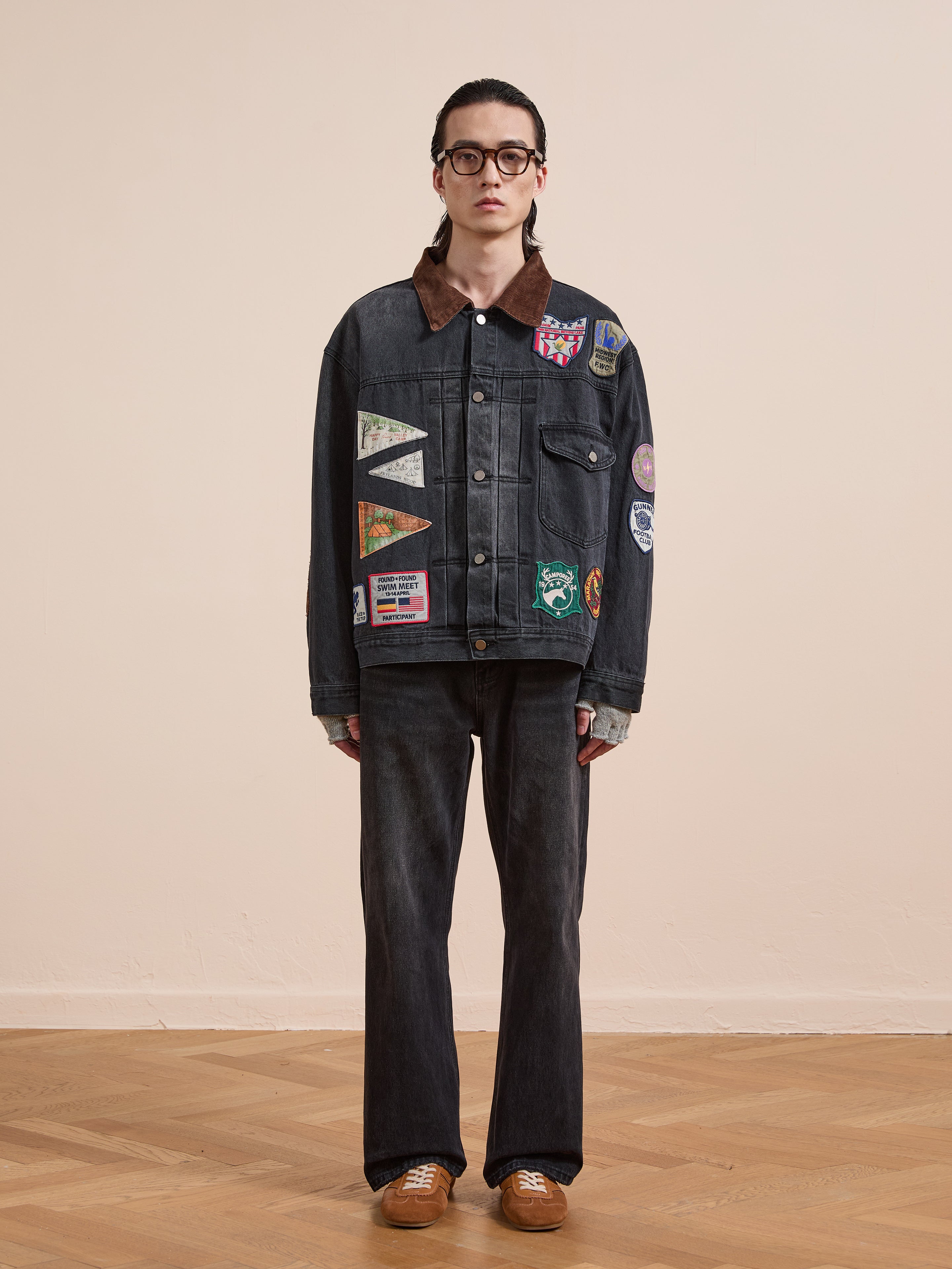 Indoors, a person wears FOUND’s Denim Patchwork Jacket with Americana patches and corduroy collar, paired with glasses, dark jeans, gray gloves, and brown shoes on a wooden floor against a plain background.