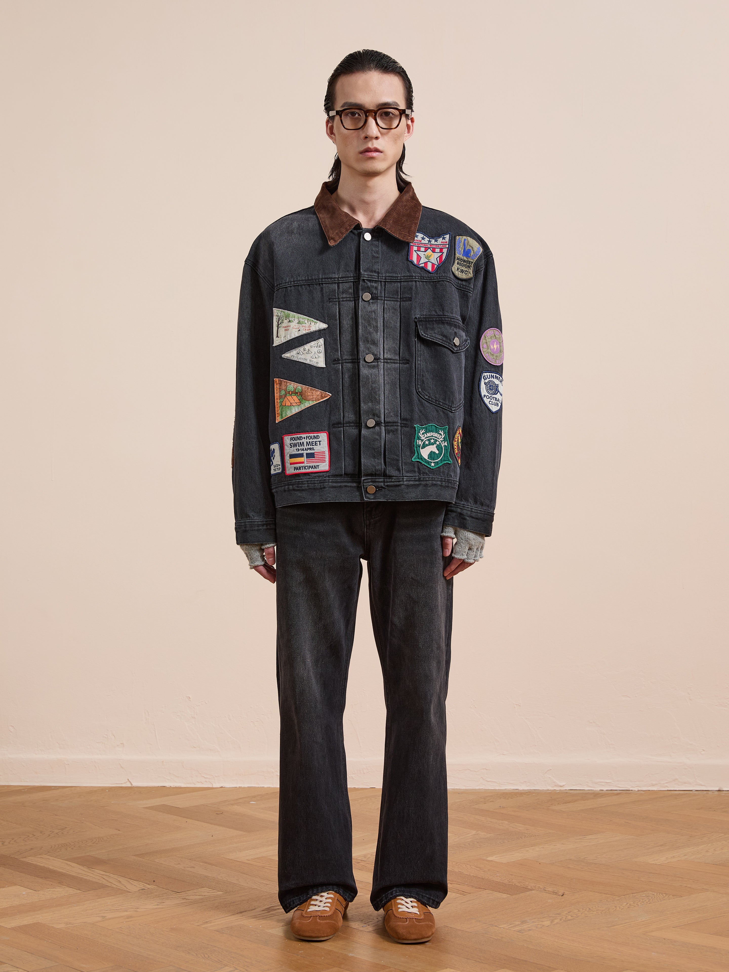 A person wearing the FOUND Denim Patchwork Jacket, black jeans, brown shoes, and glasses stands against a plain beige background on a wooden floor.